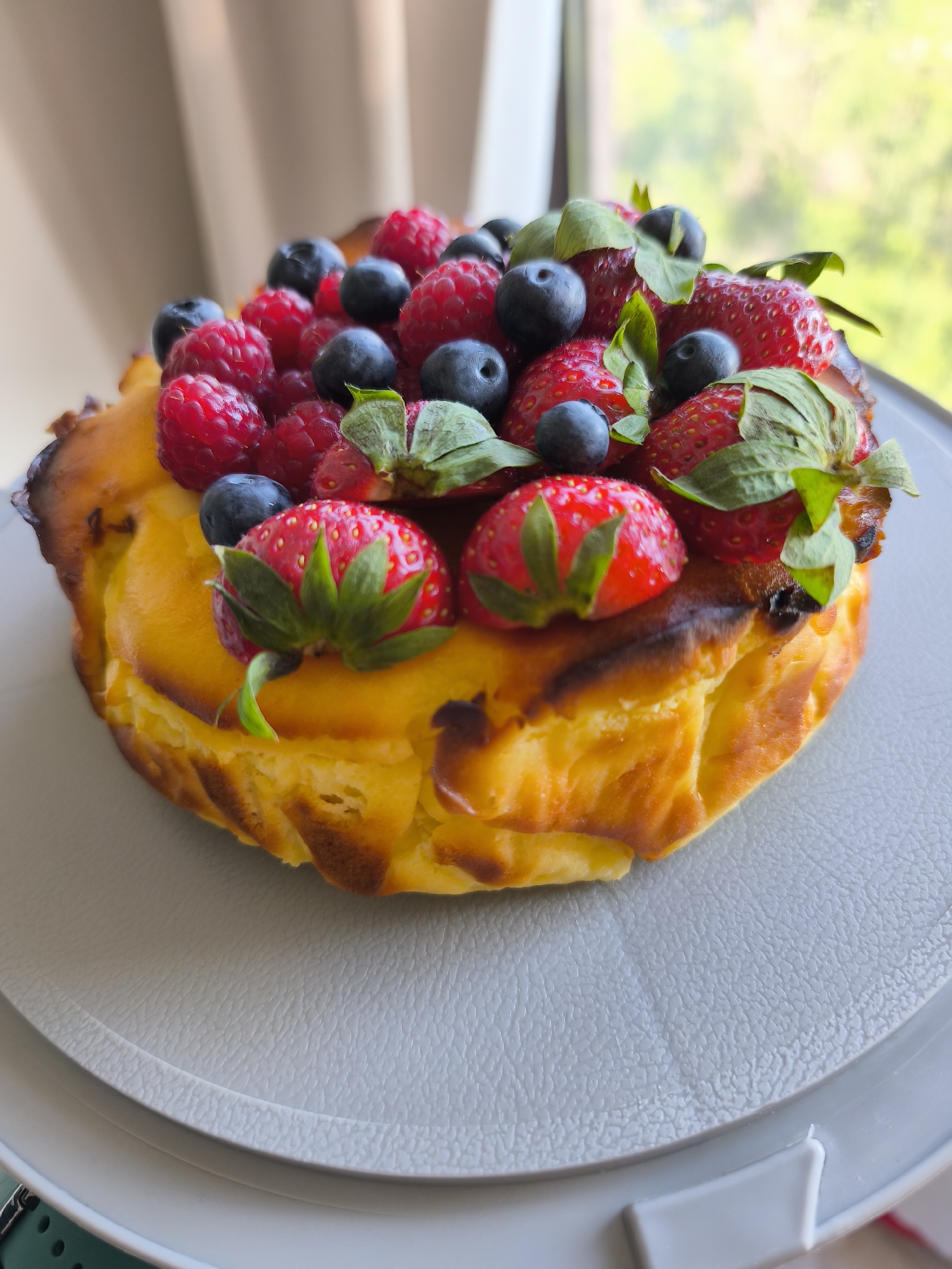A close-up shot presents a fruit-topped baked good, likely a cake or tart, centered on a textured light gray plate. The baked portion is golden-brown with a somewhat burnt crust. The topping consists of fresh strawberries, blueberries, and raspberries, arranged artfully. The window behind the cake provides soft, natural light, and a glimpse of greenery outside suggests a garden or park setting. No people are visible in the frame. This image has no identifiable ties to the city of Plovdiv, Bulgaria, or any other specific location.