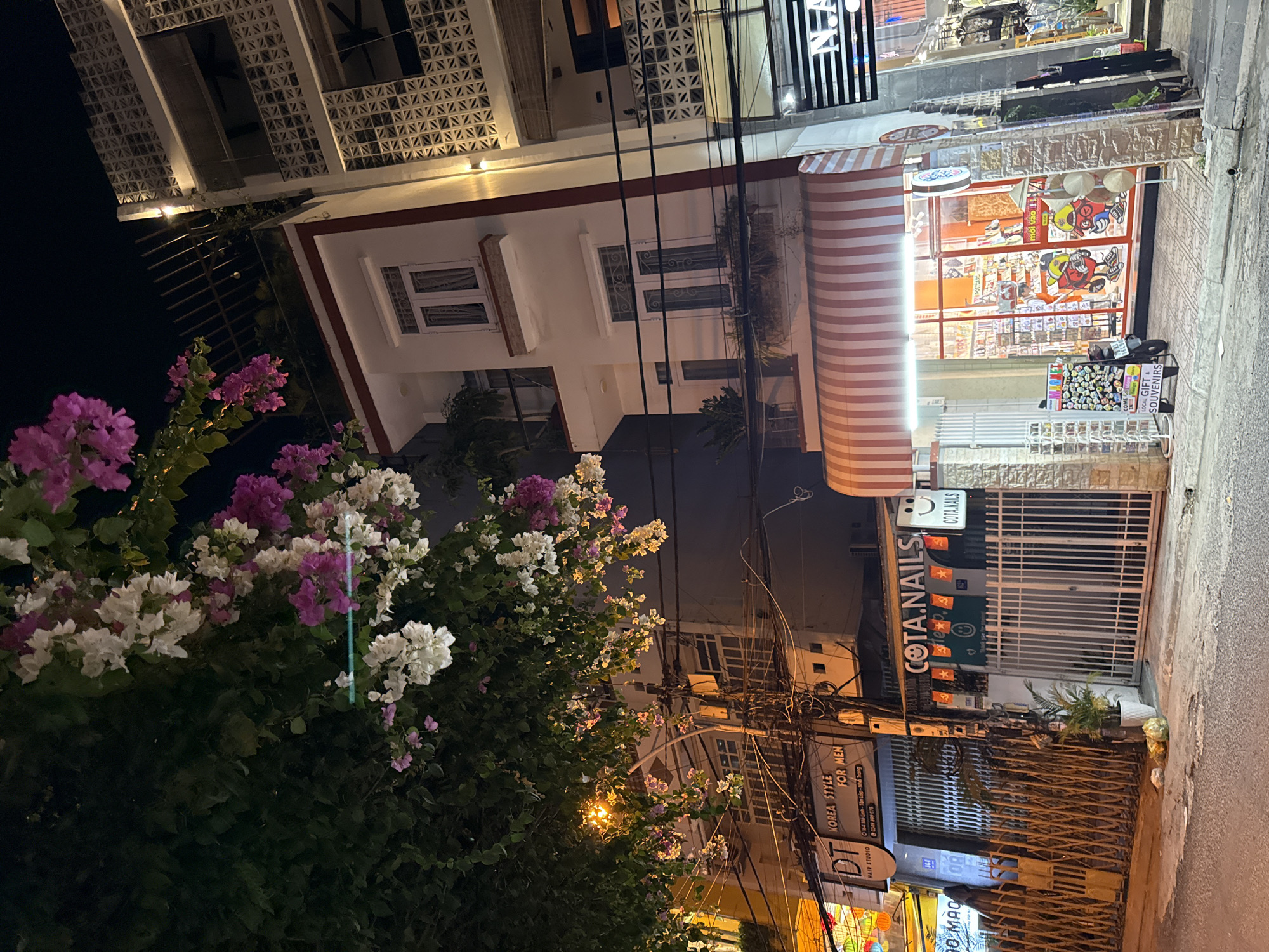 The image shows a street scene at night in Nha Trang, Vietnam. In the foreground, there is a sidewalk with several businesses. To the left, a sign reads "DT KOREA STYLE FOR MEN HAIR STUDIO". Next to it, a gated storefront displays various items for sale. Further to the right, a shop with a red and white striped awning has its windows lit up, revealing a store interior filled with merchandise. A small sign inside the window reads "mời vào", which means "come in" in Vietnamese. A motorcycle is parked on the sidewalk in front of this store. Overhead, a tree with pink and white flowers hangs over the street. Buildings with multiple stories line the street, some with decorative architectural elements. Power lines are visible crisscrossing above the street. The lighting suggests it is nighttime, with artificial lights illuminating the storefronts and the street.
