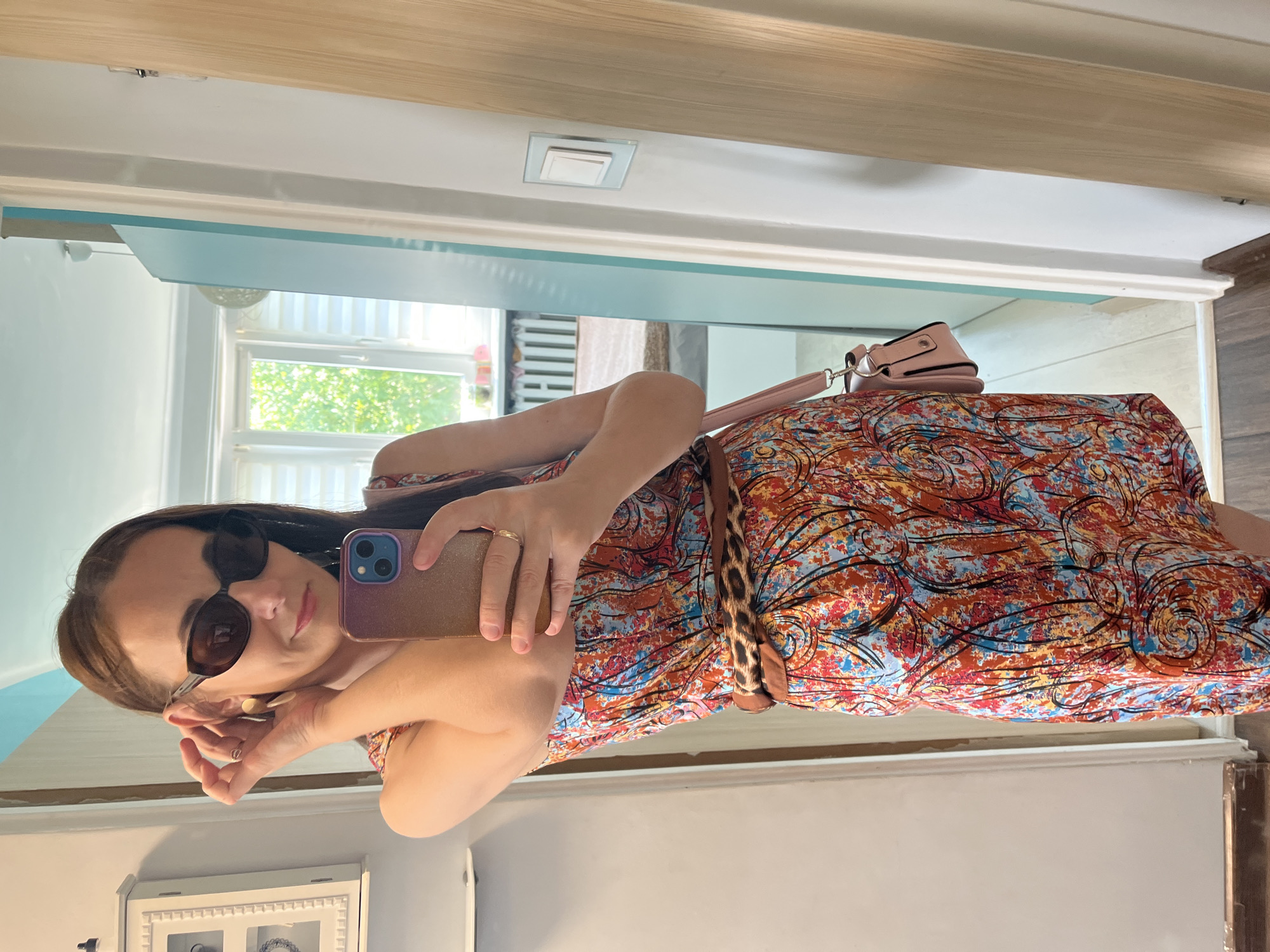 A woman is taking a selfie in a mirror. She is wearing a colorful, patterned dress with a leopard print belt and sunglasses. She has her hair down and is holding her phone in her right hand. A small pink purse is visible on her left shoulder. The background appears to be a room with white walls and a light blue accent wall. A window with blinds is visible in the background, suggesting it is daytime. The overall mood seems casual and relaxed. There is no visible text in the image.
