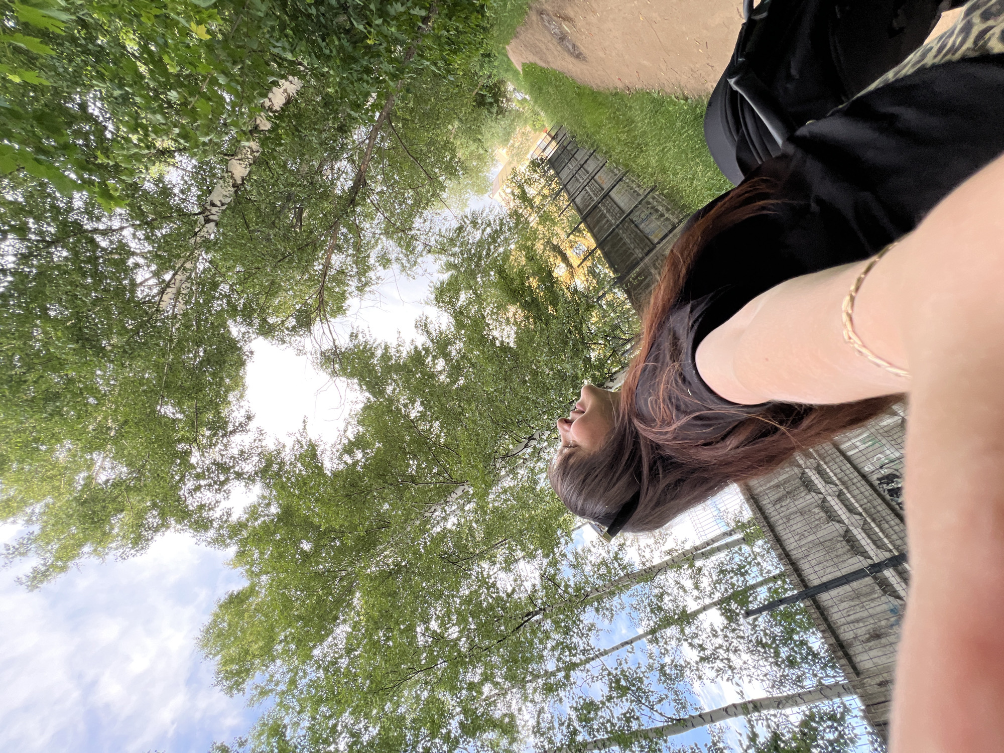 This is a low-angle, wide shot of a young woman looking up at the trees and sky, taken outdoors in Noviy, Russia. The image is oriented such that the ground is at the top of the frame and the sky is at the bottom. The woman, with long dark brown hair and wearing a black top and sunglasses, is in the foreground, her right arm and hand extended towards the camera. She is wearing a thin gold bracelet on her wrist. Her facial expression is relaxed and content.
The scene is a park or wooded area with lush green trees and a bright, cloudy sky. Sunlight filters through the leaves, creating a dappled effect. In the background, there is a dirt path, a patch of green grass, and a structure with a metal fence, possibly a greenhouse or an outdoor enclosure. The time of day appears to be daytime, likely late morning or afternoon, given the bright sunlight. The weather seems pleasant and warm.
The overall mood is peaceful and contemplative, with the woman enjoying the natural surroundings. No visible text is present in the image.