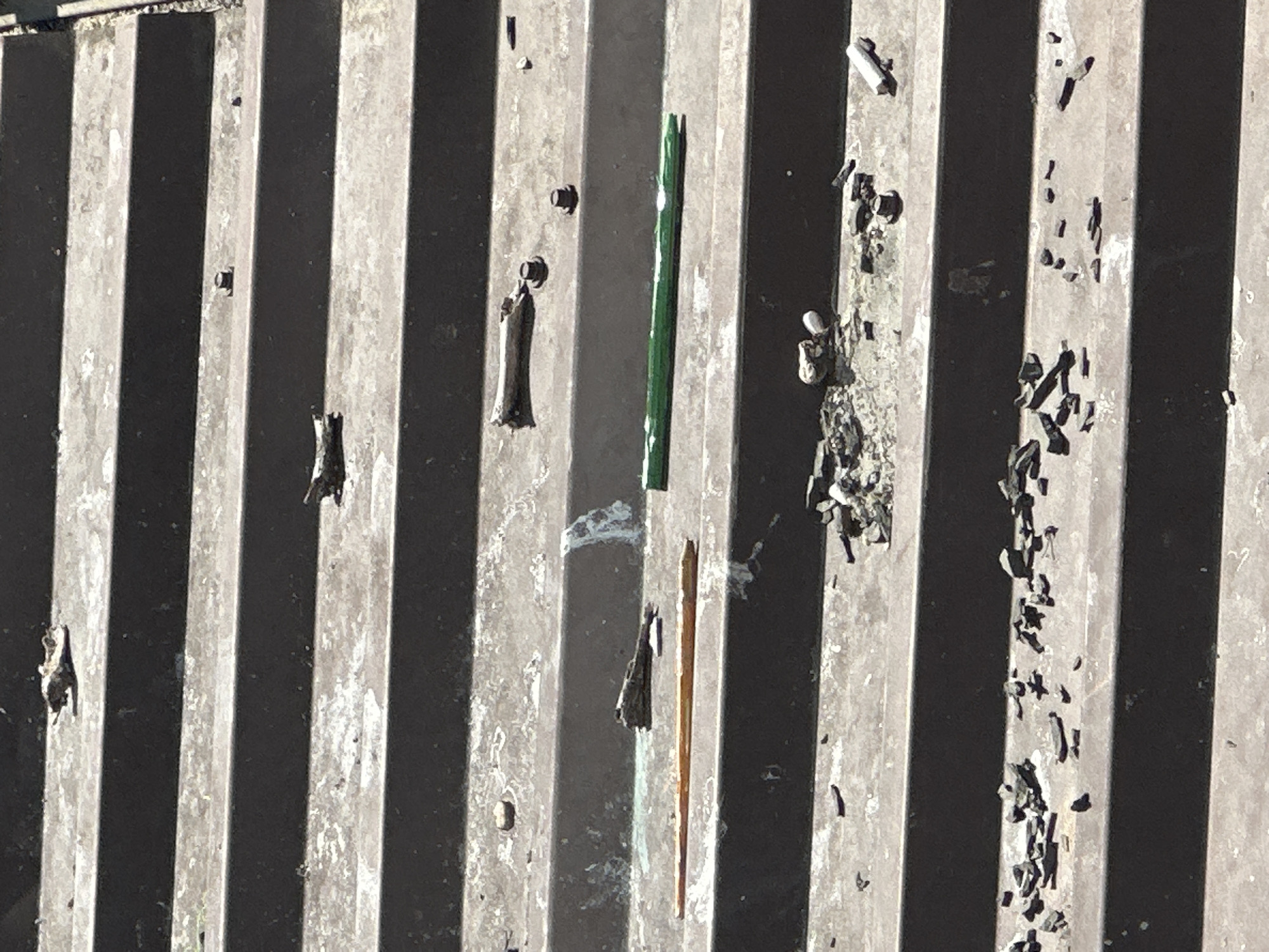 The image depicts a close-up, eye-level view of dark, weathered metal stairs, possibly in an outdoor urban setting in Sochi, Russia. The stairs are ribbed and show signs of wear and tear, with scattered debris on each step. The debris includes small pieces of wood, stones, and what appears to be fragments of glass or plastic. Notably, a long green object resembling a thick marker or crayon lies on one step, and a thin, orange object, possibly a pencil or wooden dowel, is positioned below it. Two small, dark, cylindrical objects, resembling screws or bolts, are visible on a higher step. The lighting suggests it is daytime, with shadows cast across the stairs. There are no people or animals present in the image, and no specific landmarks of Sochi are identifiable.