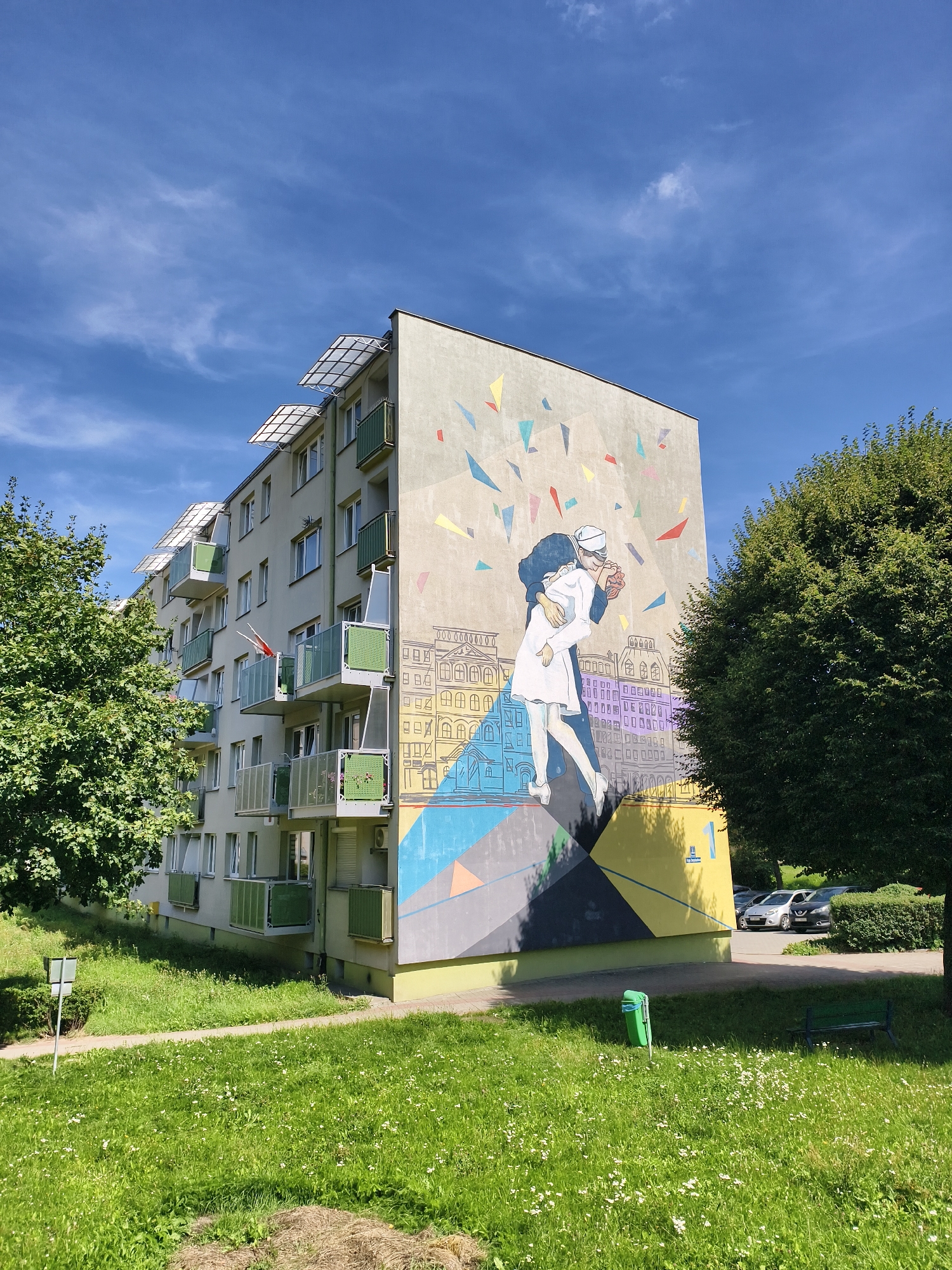 The image captures a mural on the side of a multi-story apartment building in Tczew, Poland. The mural depicts a couple embracing, with the male figure in a dark uniform and the female figure in a white dress, reminiscent of the famous "The Kiss" painting. Behind them, a stylized cityscape is rendered with various geometric shapes and a Polish flag is visible on one of the balconies. The building itself is light-colored with green and white balconies. In front of the building, there is a well-maintained lawn with scattered white wildflowers, a sidewalk, a green trash can, and a park bench. A lush green tree partially obscures the right side of the building and mural. A few cars are parked in the background, and the sky is blue with scattered white clouds.