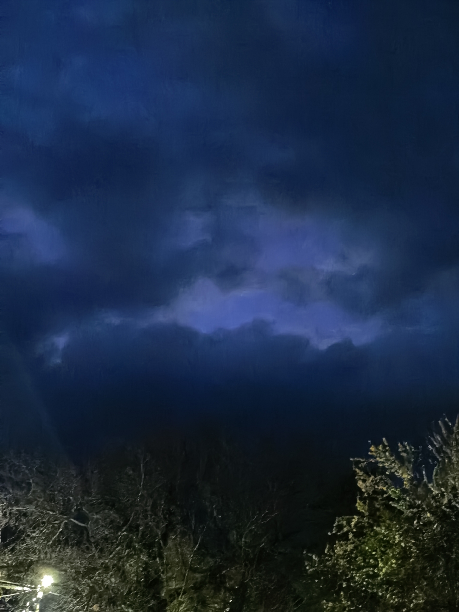 The image captures a nighttime scene in Unknown, Unknown. Dominating the upper portion of the frame are dark, heavy clouds, with a patch of lighter blue visible in the center. Below the clouds, the lower portion of the image shows vegetation, including tree branches silhouetted against the dark sky. There is a faint light source illuminating the greenery on the right side of the frame, and what appears to be a streetlight can be seen through the branches on the left. Overall, the scene is dimly lit and foreboding.
