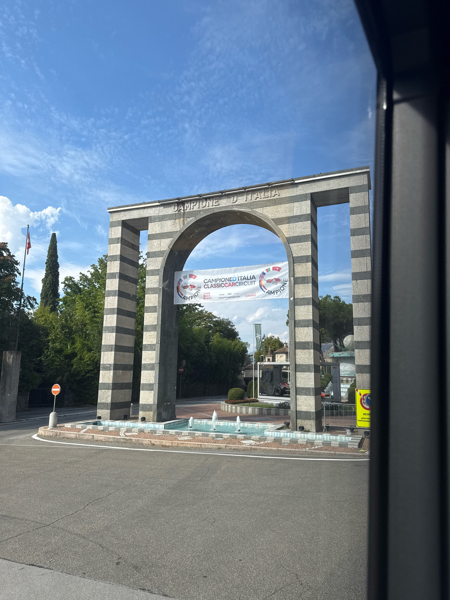 The image shows an archway in Campione d'Italia, Italy, with the words "CAMPIONE D'ITALIA" inscribed above it. A banner hangs from the arch, advertising a "CAMPIONED ITALIA CLASSICCAR CIRCUIT" event, featuring classic cars. In front of the arch is a decorative fountain. To the left, a tall, slender tree stands next to a flagpole with a Swiss flag. The background consists of lush green trees and a blue sky with scattered clouds. The foreground is a paved road with a white line marking the edge. A dark, vertical element, likely a part of a vehicle or structure, is visible on the right side of the frame. Several traffic signs are also present.