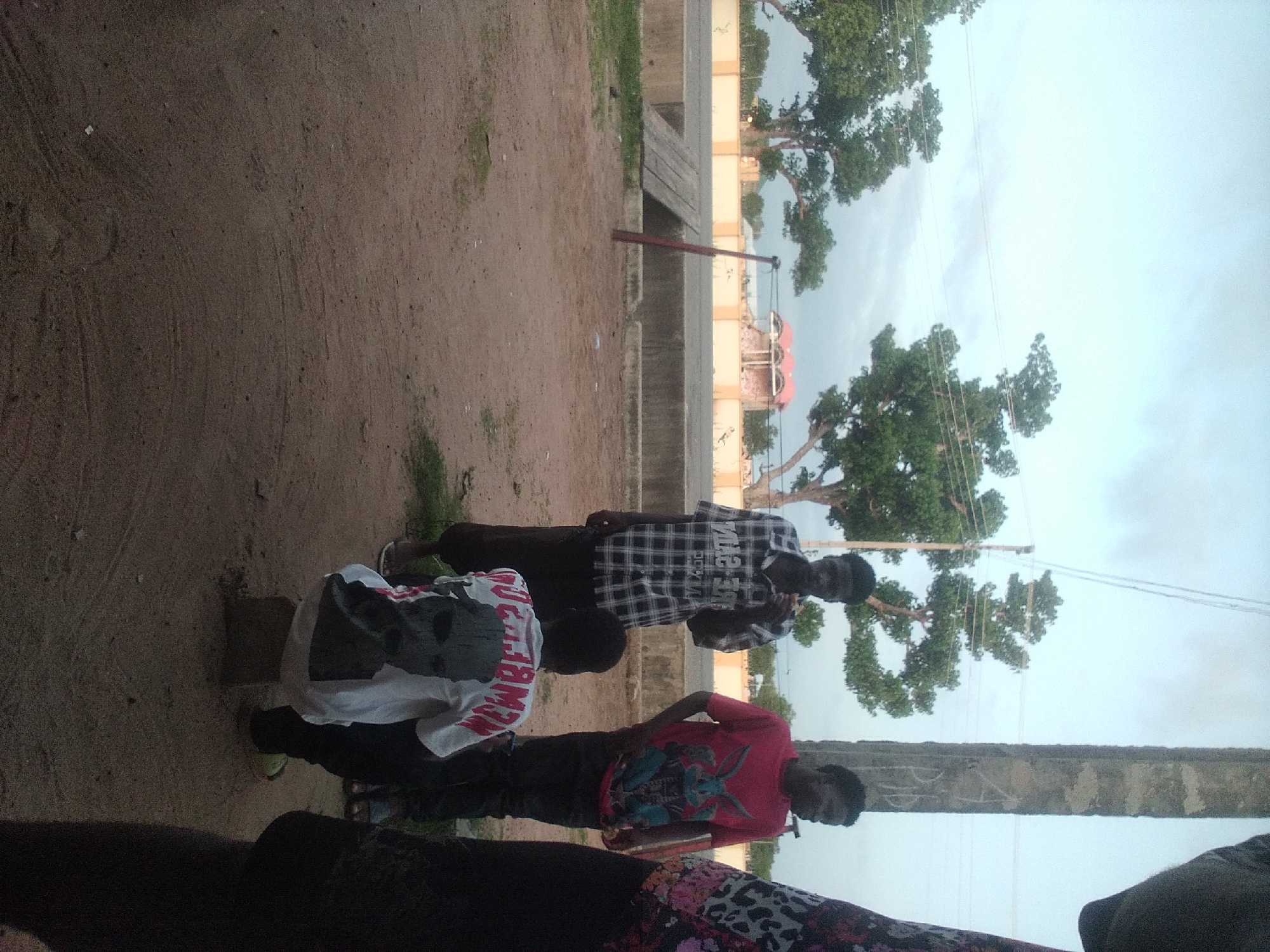 The image depicts an outdoor scene in Jalingo, Nigeria. Four individuals are visible. In the foreground, a person wearing a white t-shirt with a graphic print is seated on a small concrete block. To their left and slightly behind, a person in a checkered shirt stands, appearing to be eating. To the right of the seated person, another individual in a red t-shirt with a rabbit graphic stands. Further to the right, a portion of a person wearing a patterned garment is visible. The background shows trees, a road, and a building with an arched roof. A wooden ramp and a red pole are also present in the scene. The ground is sandy and dusty. The sky is overcast.