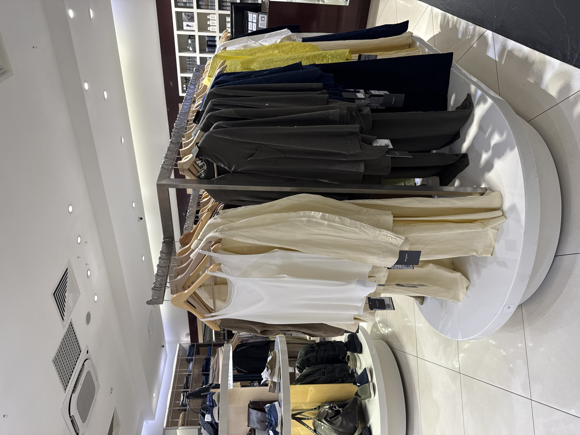 The image depicts a retail clothing store, likely a boutique, with a selection of men's or women's apparel displayed on racks and tiered shelves. The overall aesthetic is minimalist and modern, featuring white and light wood tones. In the foreground, a rack displays several garments, including a white tank top and beige linen trousers, alongside darker colored shirts and trousers. The clothing appears to be made of natural fabrics, suggesting a focus on comfort and quality.
To the left, tiered circular displays showcase shoes and handbags. A prominent brown leather tote bag is visible, along with several pairs of sneakers. The store's lighting is bright and evenly distributed, typical of a retail environment. The ceiling features air conditioning vents and recessed lighting fixtures. In the background, shelves are stocked with folded clothing and accessories, and a counter area with a computer monitor is partially visible. The floor is tiled with large, light-colored squares. There are no people visible in the image.
Based on the available visual cues, such as the modern retail design, the type of clothing displayed, and the general ambiance, this image could be located in a contemporary fashion store in Moscow, Russia. Many high-end and mid-range clothing retailers in Moscow adopt similar design elements and product selections.