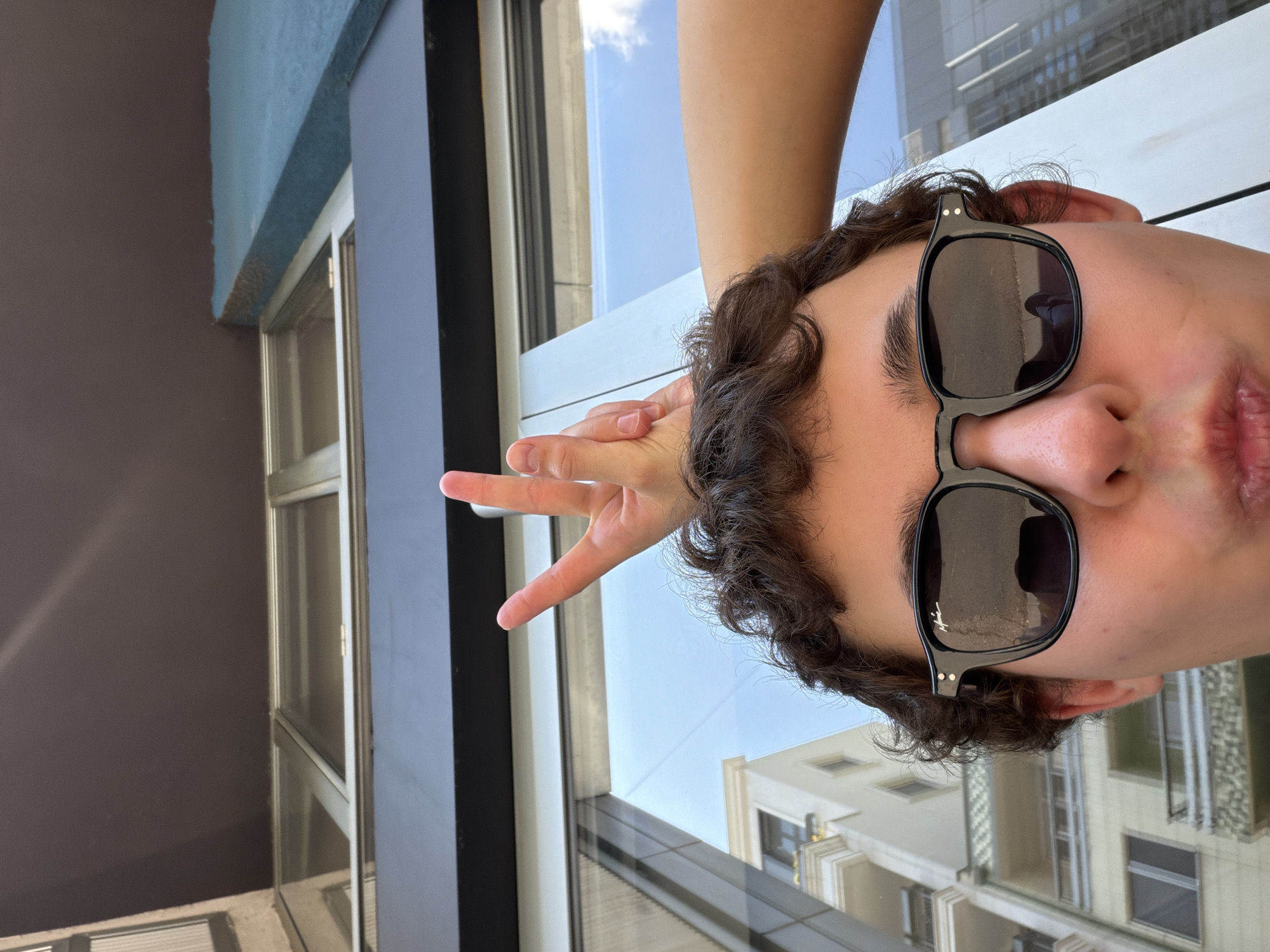 The scene is an outdoor urban setting in Vracar Urban Municipality, Serbia. In the foreground, the lower half of a person's face is visible, featuring a nose, a slightly puckered mouth, and a chin. The person wears black-framed sunglasses that reflect the surrounding urban landscape and sky. Dark, curly hair frames the face. Above the person's head, an arm extends from the right, with a hand positioned over the head. This hand is performing a "peace" sign, with the index and middle fingers extended upwards in a V-shape. The background primarily consists of a dark, solid overhead structure transitioning into large reflective windows. These windows and the sunglasses reflect various buildings, including a light-colored multi-story building with balconies on the left, and a structure with vertical lines on the right, all under a clear blue sky with some cloud formations.