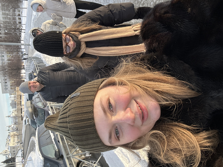 The image depicts a snowy urban scene featuring a group of six individuals outdoors under clear, sunny winter conditions. The foreground is dominated by a young blonde female, aged approximately 20-30, wearing an olive green ribbed beanie and a dark fur coat, smiling directly at the camera, suggesting a selfie. Braces are visible on her upper teeth. To her right and slightly behind, a second female, aged approximately 20-30, wears a black ribbed beanie, brown-rimmed glasses, a beige ribbed scarf, and a black puffer jacket; she looks downwards. Further back, a male, aged approximately 40-50, wears a light gray ribbed beanie and a black puffer jacket, smiling towards the camera's general direction. Behind them, two additional individuals, one male in a light-colored hooded jacket and beanie, and another female in a light-colored hooded jacket and beanie, are partially visible, both appearing to be walking. A sixth individual, obscured, is also present behind the second female. The ground is covered in a uniform layer of snow.
In the midground, a row of parked vehicles, including SUVs and wagons, some equipped with roof cargo boxes, are visible behind a dark metal barrier or fence. These vehicles are snow-covered.
The background features a prominent architectural complex consisting of white buildings with dome-like structures, characteristic of the Kazan Kremlin in Kazan, Russia. One building, possibly a cathedral or mosque, has multiple large, light-colored domes, some with golden accents, suggesting religious architecture. The complex is situated on an elevated landmass. Distant, less distinct urban buildings and leafless trees are also visible. The scene appears to be along an embankment or pathway overlooking the Kremlin.