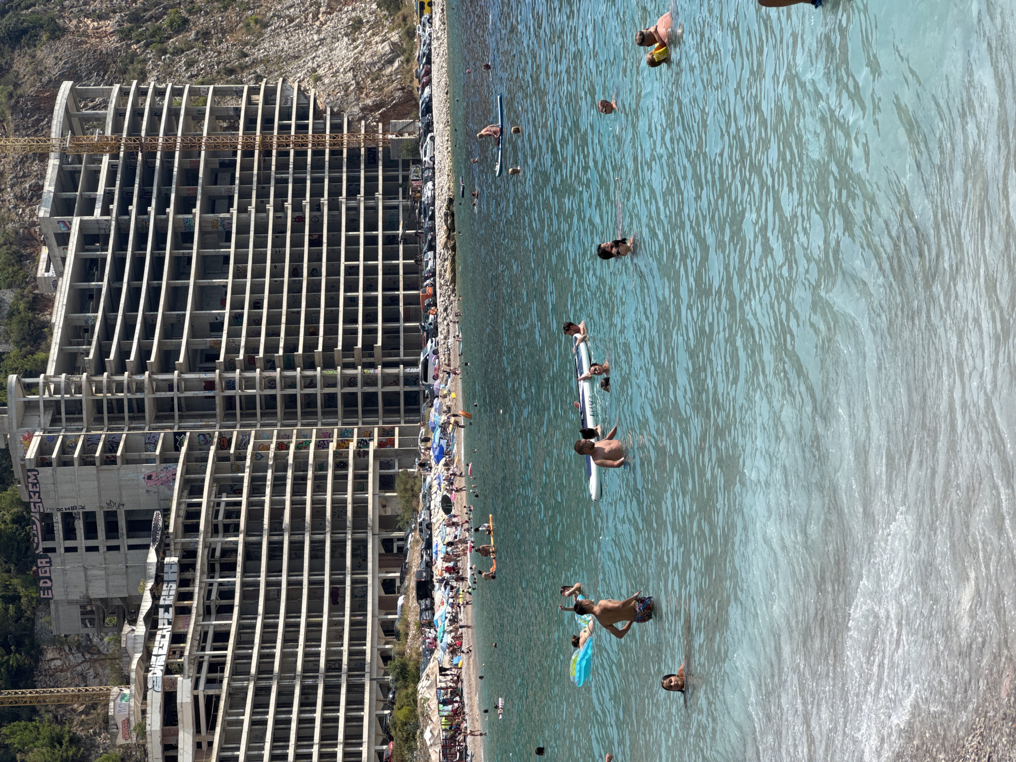 In Mirište, Montenegro, the scene depicts a busy beach area with a large, unfinished concrete building in the background. The building is multi-story and appears to be under construction, with exposed rebar and numerous windows. Graffiti is visible on its exterior. In front of the building, a rocky shoreline is lined with parked cars and umbrellas, indicating a public beach. The water is a clear, turquoise blue, and filled with numerous people swimming, sunbathing on inflatables, and paddleboarding. Several individuals are seen interacting in the water, with some swimming close to shore and others further out. The overall atmosphere suggests a recreational summer day at the beach.