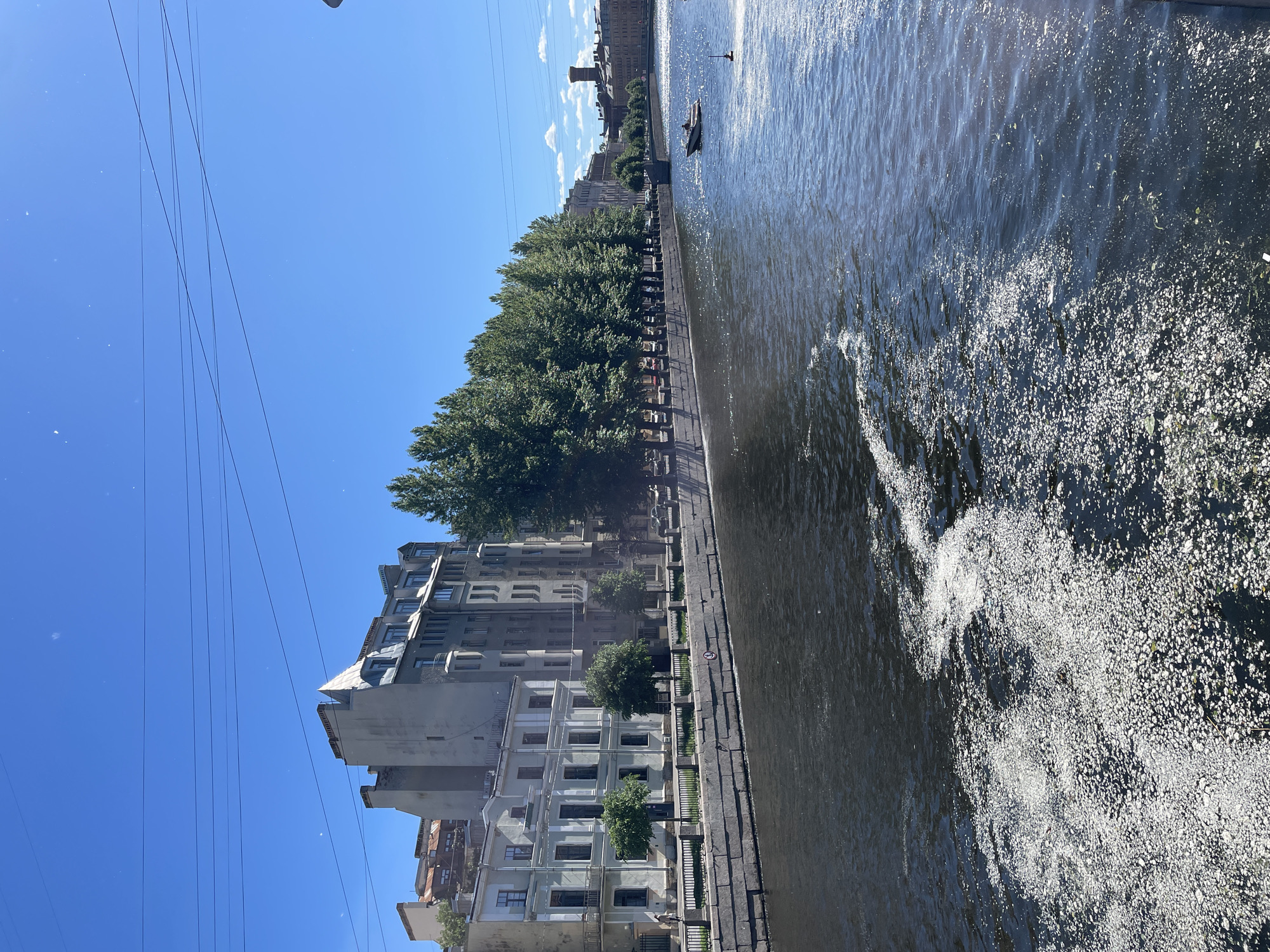 The image depicts a sunlit scene along a canal in Saint Petersburg, Russia. The perspective is from a boat on the water, looking towards the city embankment. Tall, light-colored buildings with numerous windows line the canal, interspersed with lush green trees. A paved walkway runs alongside the canal, bordered by a low fence.
In the distance, a silhouette of a person in a small boat is visible on the water, suggesting a leisurely activity. The water itself is dark and choppy, with white foam and debris concentrated on the right side of the frame, indicating movement or a disturbance. The sky is a clear, bright blue, with a few wispy clouds. Power lines stretch across the upper left portion of the image. The overall impression is a bright, clear day, likely in the late morning or early afternoon due to the strong sunlight and shadows. The architecture suggests a historic European city. No visible text is present in the image.