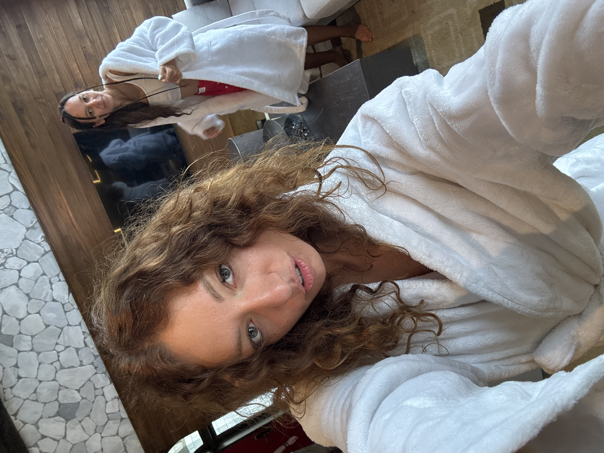 The image shows a selfie of a woman with curly hair wearing a white bathrobe. In the background, another person is also wearing a white bathrobe and lying on a surface. A dark table and a grey couch are visible in the background. The location appears to be indoors, possibly a hotel room or spa. The setting resembles modern interior design with elements such as wood paneling, stone flooring, and a flat-screen TV. There is no clear evidence to confirm that this photo was taken in Cat Cat Village, Vietnam.
