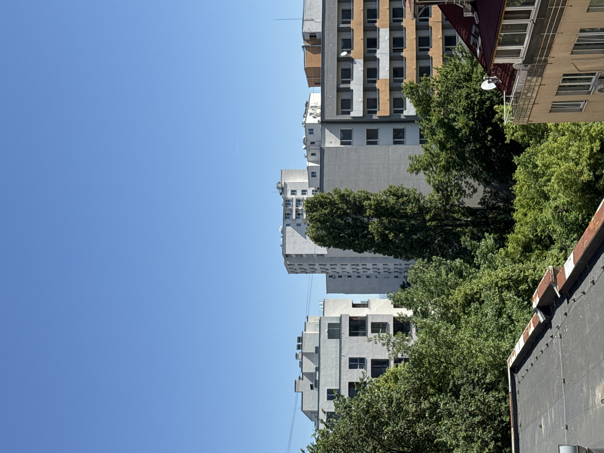 The image shows a view of several buildings and green trees against a clear, blue sky. The buildings are of varying heights, with a mix of architectural styles and facade colors, ranging from gray to beige and featuring repetitive window patterns. Lush green trees are present, partially obscuring the buildings and providing a contrast to the urban landscape. The location is likely in the city of Sofia, Bulgaria.