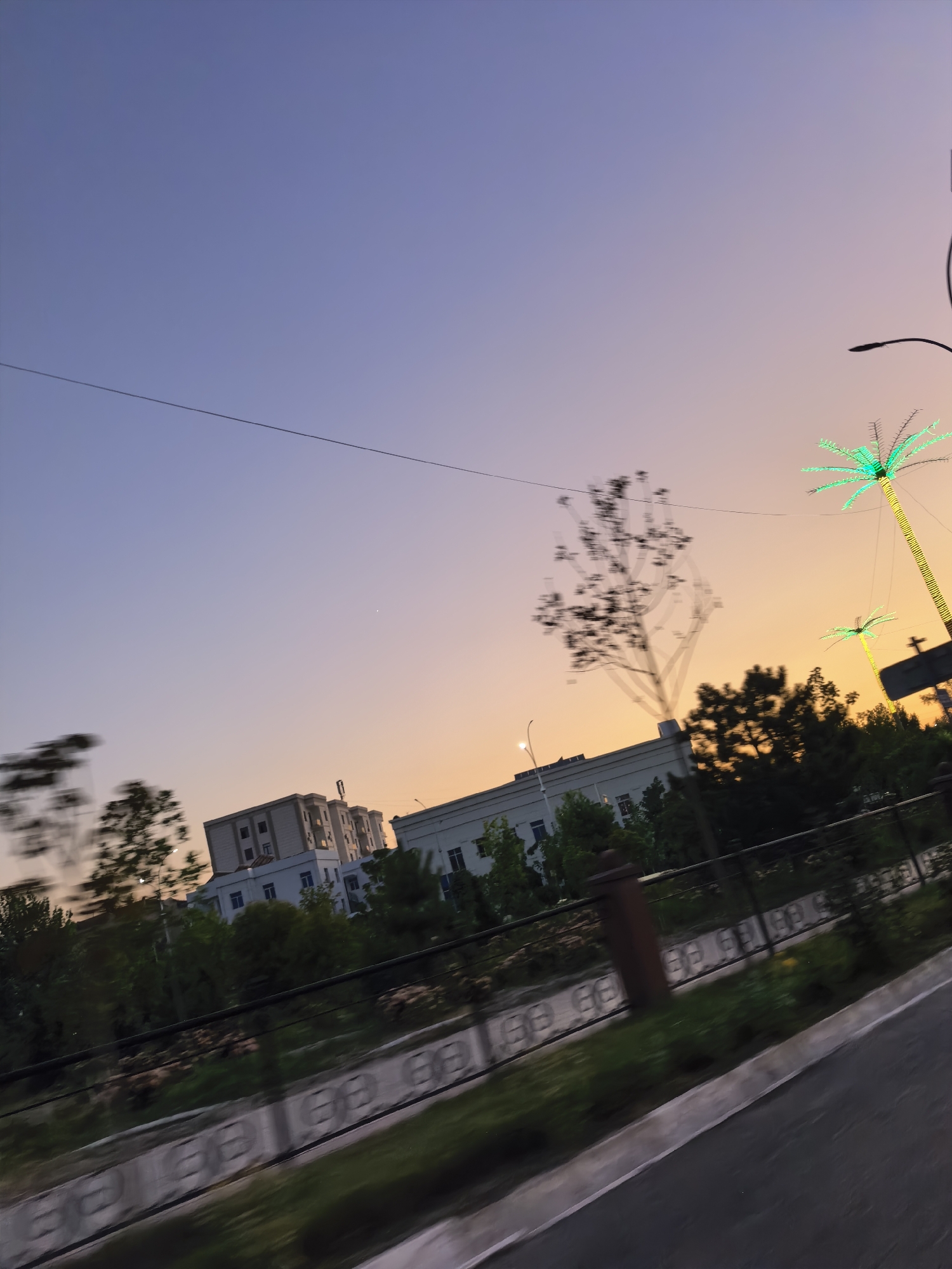 The image depicts a city street scene in Fergana City, Uzbekistan, during twilight. The sky transitions from a soft purple overhead to a warm orange hue near the horizon. Buildings with multiple stories, appearing modern and white, are visible in the background. Green foliage and trees line the street, interspersed with a black metal fence. Decorative, illuminated palm trees, lit with green lights, stand out against the darkening sky, suggesting a festive or nighttime ambiance. A utility wire stretches across the upper portion of the frame. The perspective indicates the photo was likely taken from a moving vehicle, evidenced by the motion blur in the foreground and parts of the midground. The overall impression is that of an urban environment transitioning into evening.
