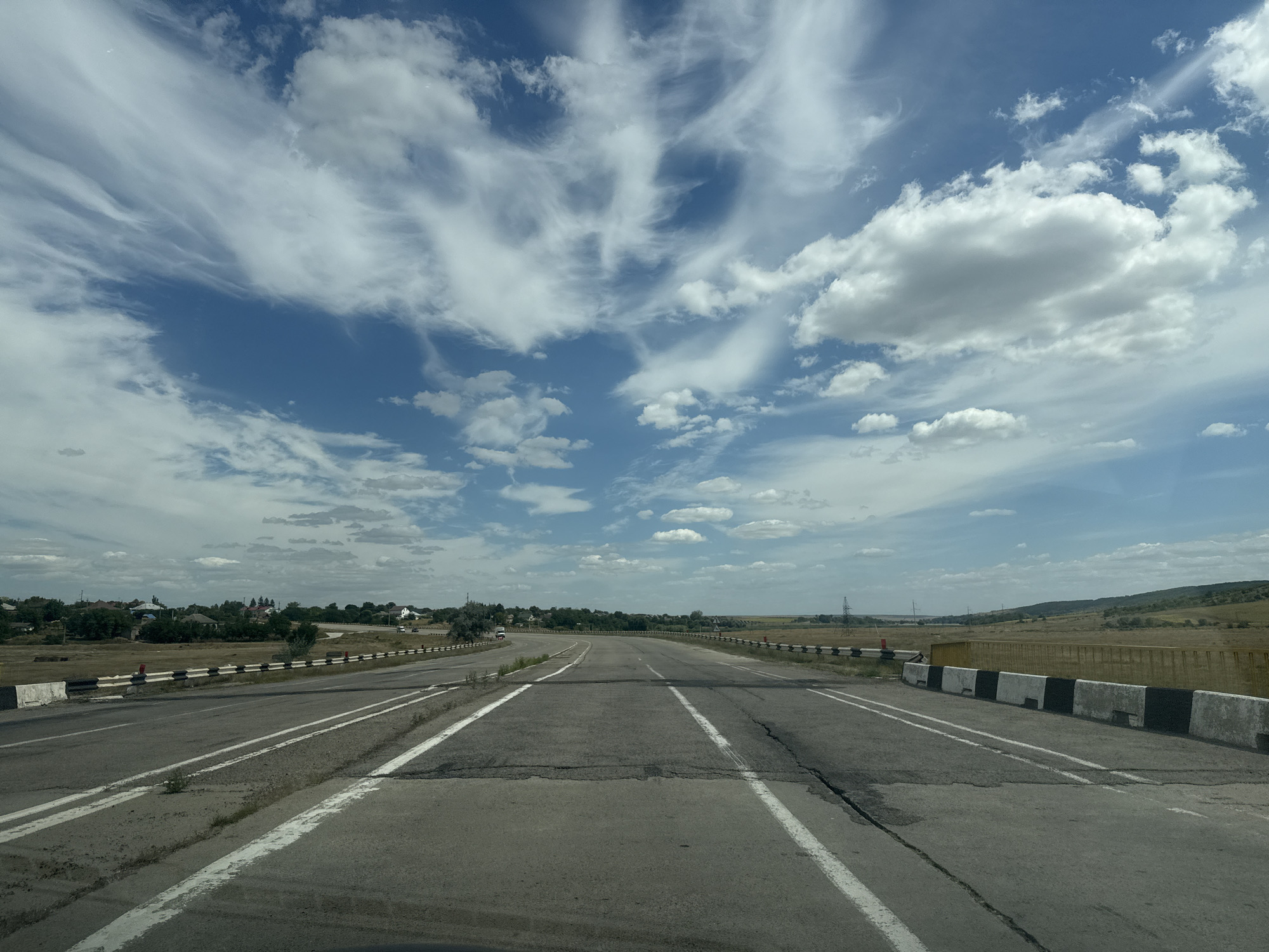 The image shows an empty, cracked asphalt road with white lane markings, leading into the distance. The road is bordered by concrete barriers with black and white stripes. On the left side of the road, there are some houses and trees in the background, suggesting a semi-rural or suburban setting. The sky is blue with scattered white clouds, some of which are wispy cirrus clouds. The overall impression is of a highway or major road passing through a landscape with scattered human settlements. The specific location is identified as the city of Razeni, Moldova.