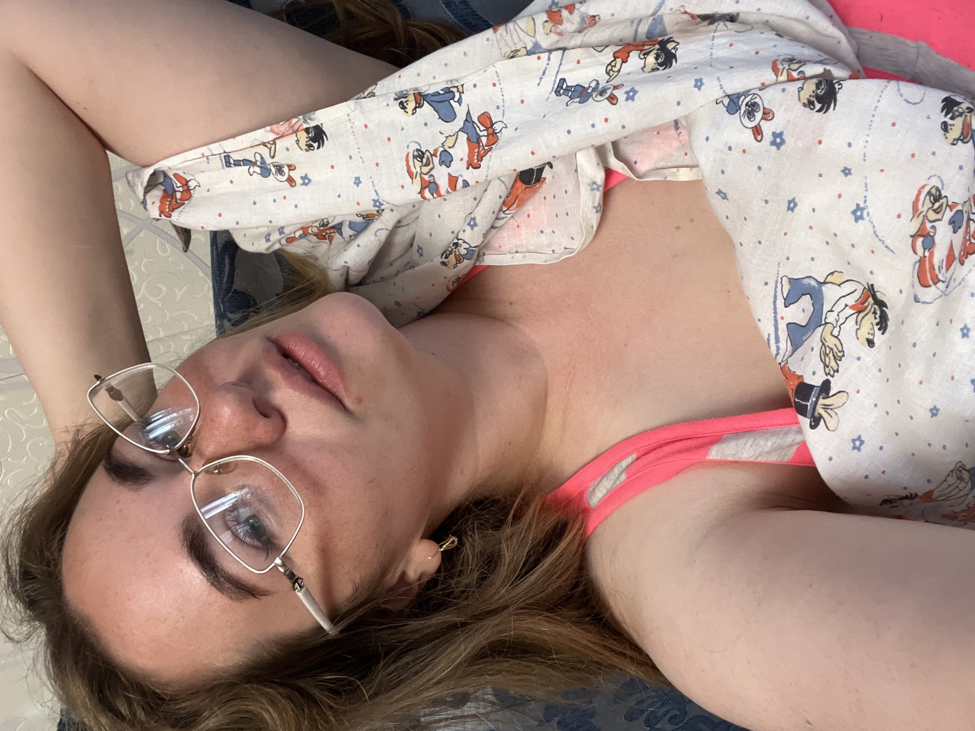 The image presents a close-up, slightly tilted perspective of a young woman lying down indoors, likely in a residential setting in Chaikovsky, Russia. Her head is positioned towards the bottom of the frame, and she is looking directly into the camera through a pair of gold-rimmed eyeglasses. Her long, light brown hair cascades around her head and shoulders. Her facial expression is neutral, with her lips slightly parted, suggesting a moment of repose or contemplation.
She is wearing a bright pink top or bra strap, partially visible beneath a light-colored blanket or sheet. This covering features a whimsical pattern of cartoon characters resembling classic animated figures like Mickey Mouse, Donald Duck, and Goofy, along with scattered blue stars and red dots. A small, simple earring is visible in her right ear. The background consists of a textured, light-toned surface, possibly a bedspread or upholstery, reinforcing the impression of an indoor, domestic environment. There are no specific cues for the time of day or outdoor weather. No visible text is present in the image. The overall scene conveys a casual and relaxed atmosphere.
