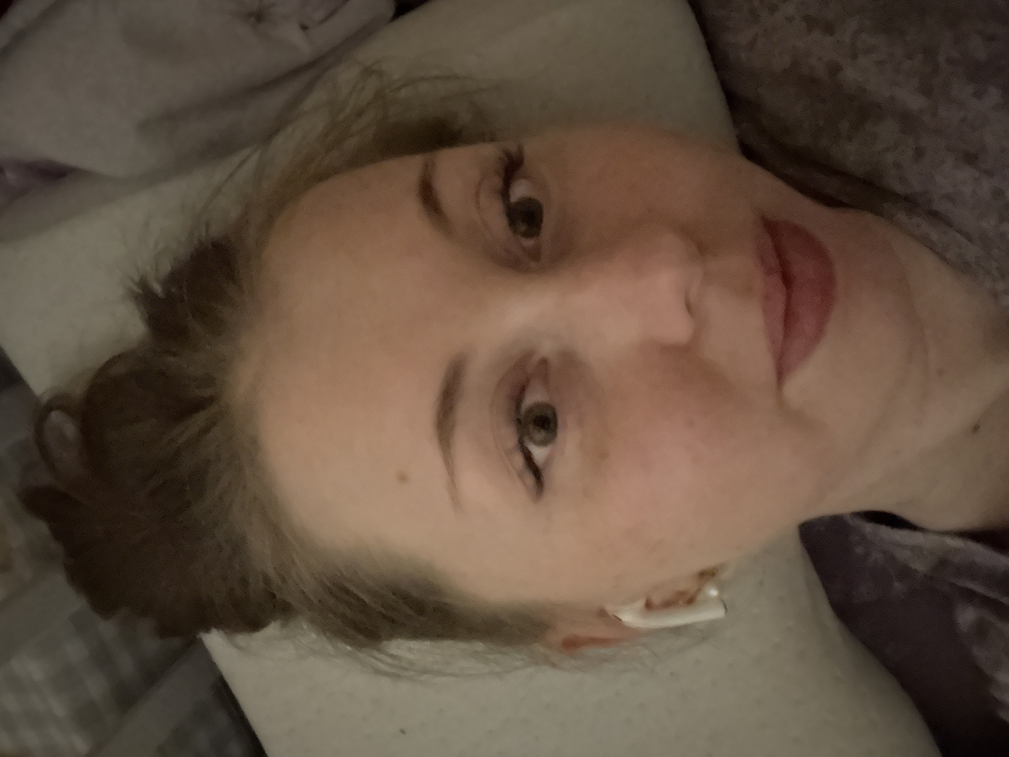 The image depicts a person's face in close-up. The person has fair skin, freckles, and brown hair. They are lying on a white pillow with a gray blanket nearby. An earbud is visible in the person's right ear. The location is Unknown, Unknown.