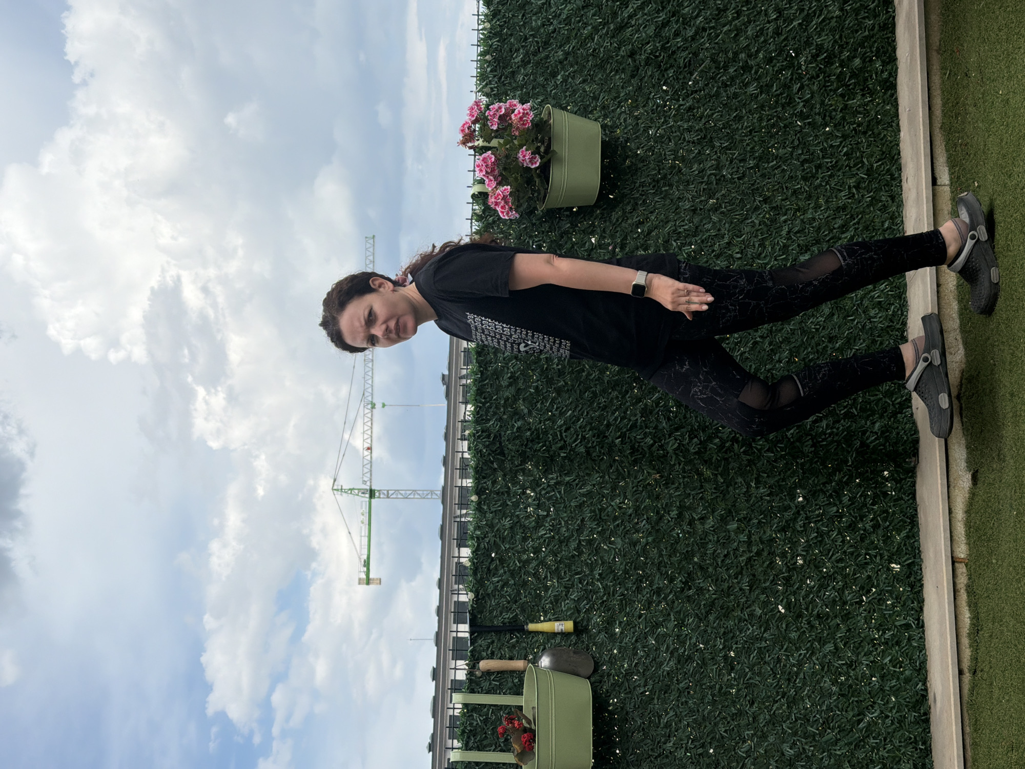A woman with dark hair, wearing a black t-shirt and black patterned leggings, stands on a green, artificial grass surface. She is looking towards the camera with a neutral expression. She is wearing black Crocs-style shoes. To her left, a green planter box with pink flowers is mounted on a vertical green hedge. Behind the hedge, a grey railing runs horizontally across the frame, and beyond that, a large green and white construction crane is visible against a cloudy blue sky. In the foreground, on the artificial grass, are garden tools including a trowel and a watering can. The scene appears to be outdoors, possibly on a balcony or rooftop, given the presence of the construction crane and the urban backdrop. The weather appears partly cloudy. No text is visible in the image.