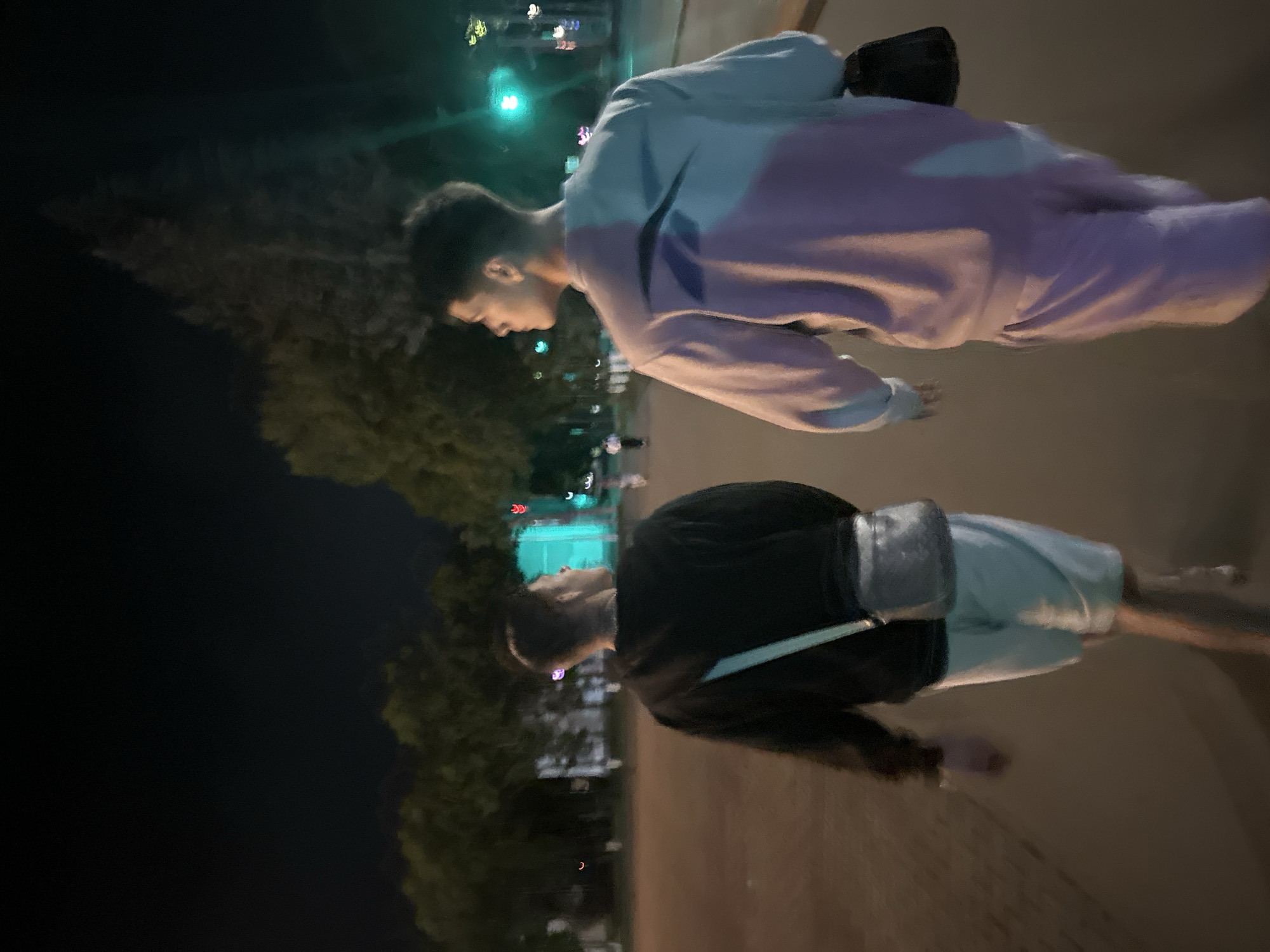 Two young men are walking away from the camera on a paved path at night. The man on the left is wearing a black sweatshirt, light-colored shorts, and a crossbody bag with a light blue strap. The man on the right is wearing a light purple and blue sweatshirt and light purple shorts. Both men are looking to their right. The background shows trees, streetlights, and buildings. The scene is likely outdoors in a city. The image quality is low due to the darkness and possibly camera shake.