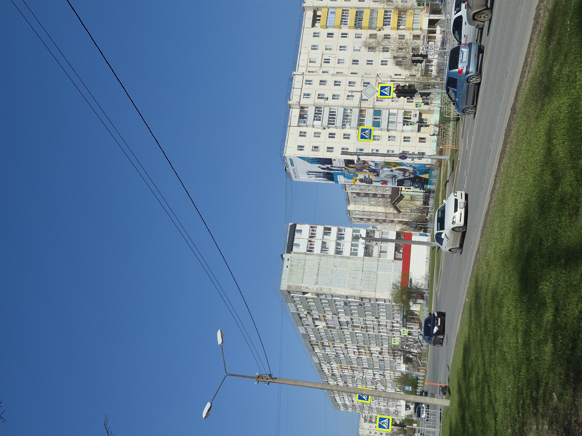 The image shows a street in Tobolsk, Russia, with multi-story apartment buildings on either side. Several cars are parked or driving on the road. Pedestrian crossing signs are visible. Overhead power lines stretch across the sky. The sky is blue. Green grass lines the side of the road. Street lamps are present. A large mural decorates one of the buildings.