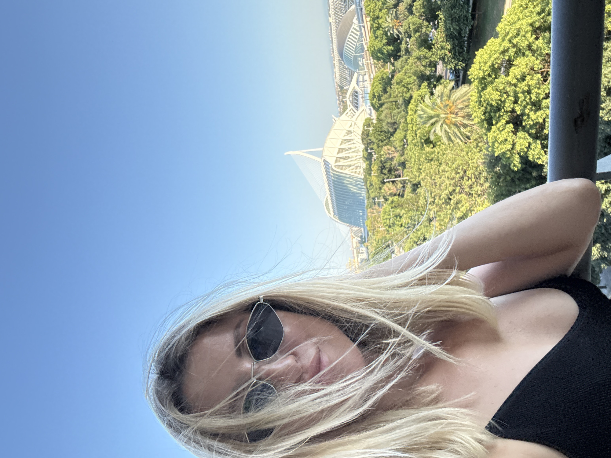 A woman with blonde hair and sunglasses is taking a selfie on a balcony with a clear blue sky. In the background, the City of Arts and Sciences in València, Spain, is visible, including the Hemisfèric and the Oceanogràfic's L'Àgora. Lush green trees surround the architectural complex, and a railing is in the foreground.