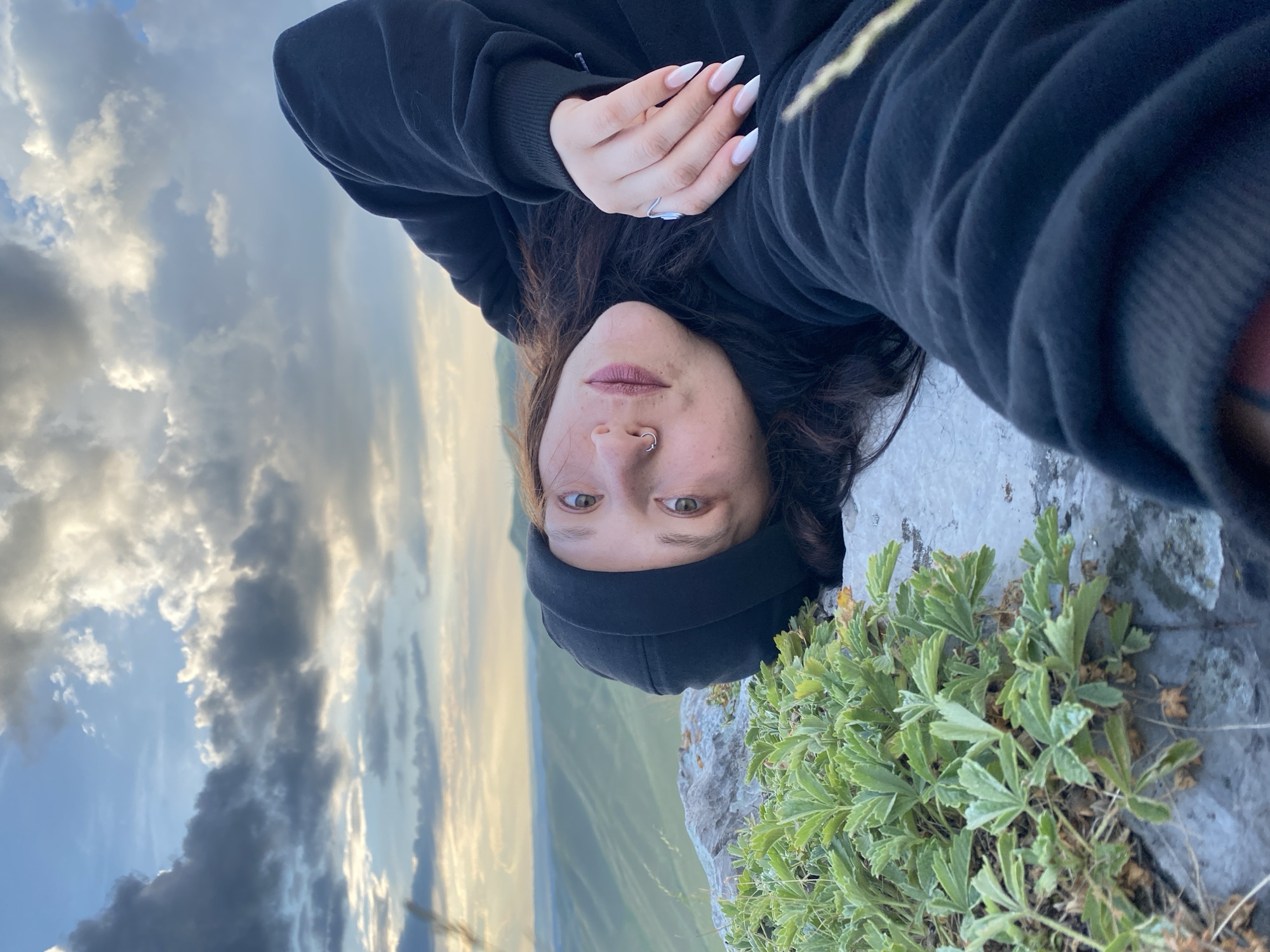 A woman lies on a rocky surface with green plants growing from the rocks. She is wearing a black hooded sweatshirt and a black beanie. Her head is turned towards the camera, and she is looking at the viewer. Her right hand is resting on her chest, and she has long, pointed fingernails painted white. In the background, there are rolling green hills under a cloudy sky. The location is the area around Абзаново, Russia.