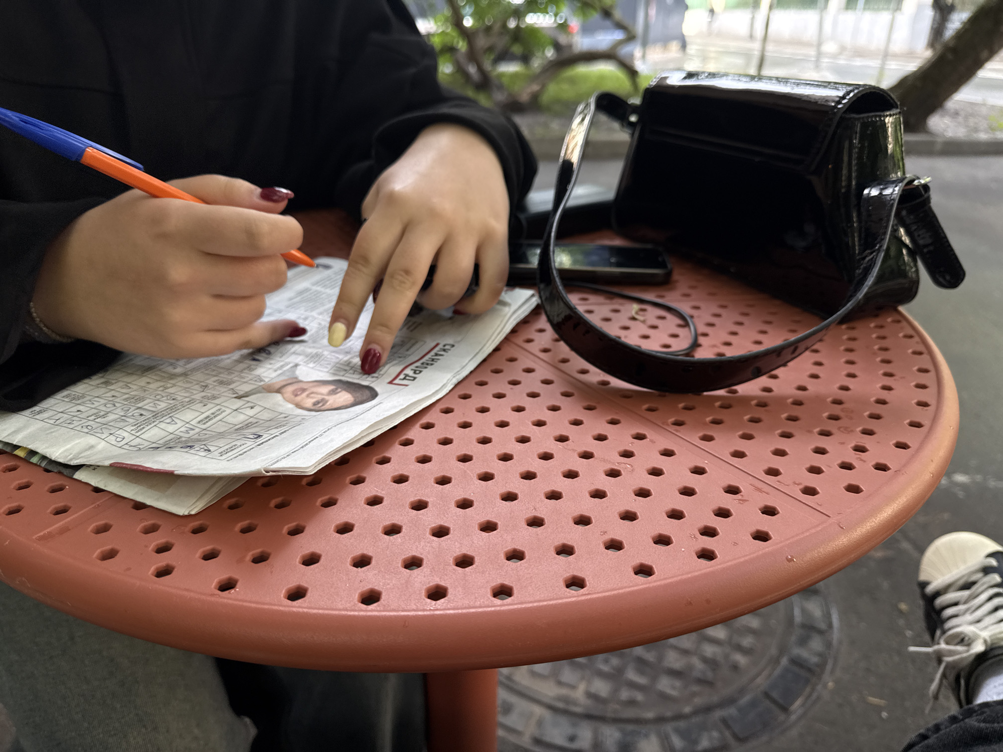 In Moscow, Russia, on a round, perforated, coral-colored table, a person wearing a black sweatshirt is writing on a newspaper with an orange and blue pen. The newspaper contains text and a photograph of a woman. Beside the newspaper, a black, shiny, patent leather handbag with a strap is placed on the table. A smartphone is also visible on the table, next to the handbag. In the background, out-of-focus greenery and trees suggest an outdoor setting. To the right, a pair of black and white sneakers with white laces are visible on the ground. The scene appears to be an outdoor cafe or public seating area.