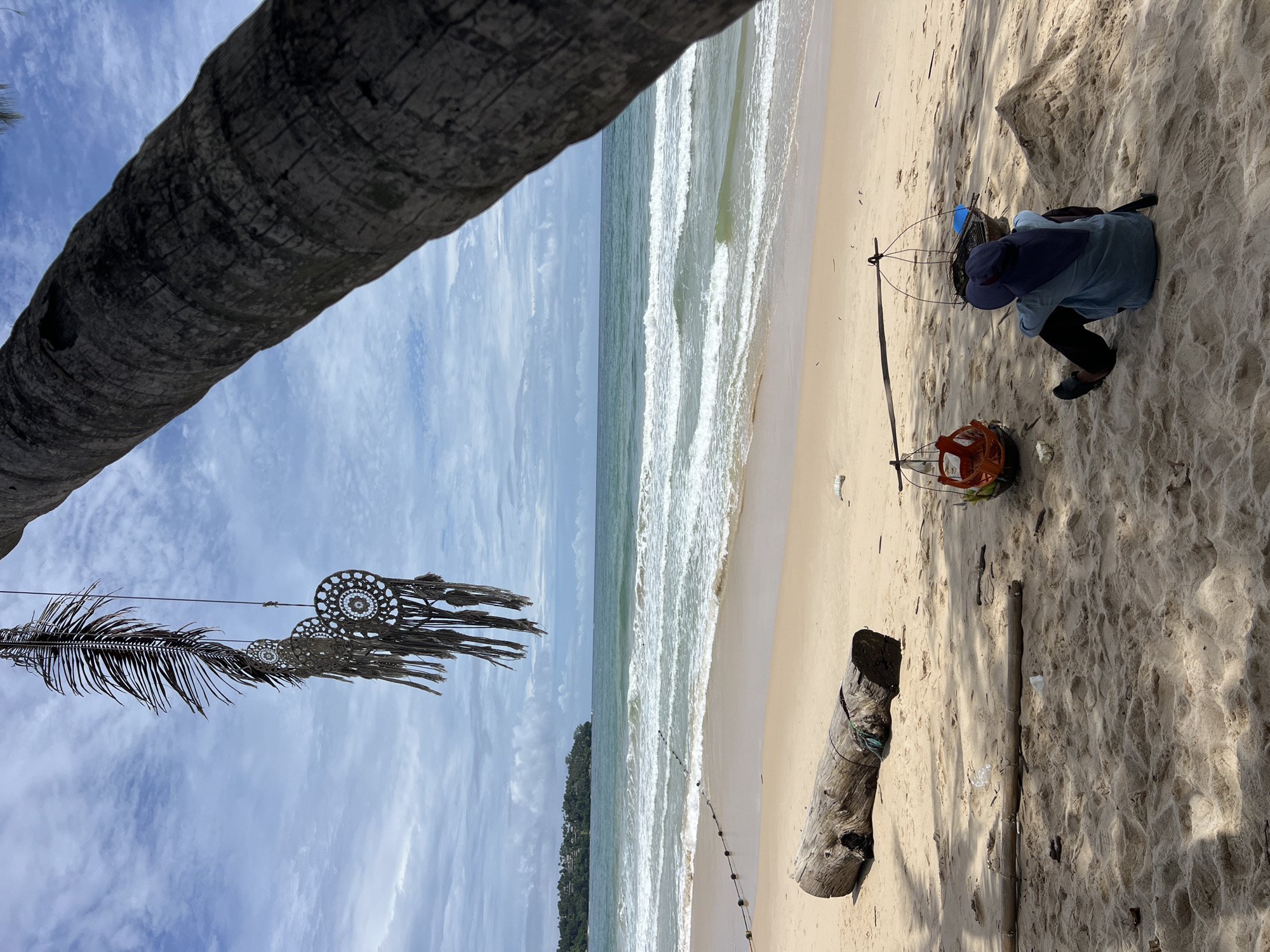 A person sits on a sandy beach, facing away from the camera, wearing a blue shirt and a dark blue hat. They are seated beside a traditional carrying pole balanced on their shoulders, with two baskets hanging from either end. One basket, made of woven material, contains an orange object, possibly food or goods. The other basket holds what appears to be a cooking grill and a blue container. To the left of the person, a large piece of driftwood lies on the sand. In the background, the ocean stretches to the horizon, with waves breaking on the shore. The sky is partly cloudy. A palm tree trunk dominates the right foreground, and decorative dreamcatchers made of natural materials hang from a branch in the upper left. The scene appears to be a quiet, tropical beach.