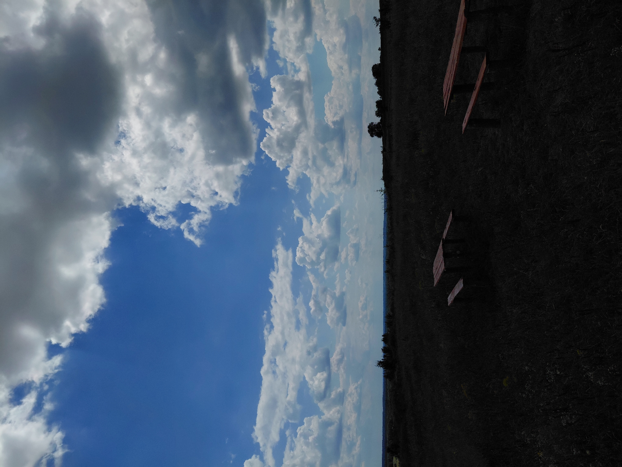 This image depicts a natural outdoor scene, likely a field or park, located in Ustyvytsia, Ukraine. The upper portion of the image is dominated by a dynamic sky with both bright blue patches and numerous white and grey clouds. The clouds vary in density, with some appearing fluffy and scattered, while others are larger and more ominous.
The lower half of the image features a dark, low-lying field with sparse vegetation. In the foreground, two wooden picnic tables with attached benches are visible. These structures are made of reddish-brown wood and are positioned in the field. One table is located towards the left side of the frame, and the other is situated further to the right and slightly closer to the viewer. The rest of the field is dark, obscuring finer details of the ground cover and any potential flora.
In the distance, a line of dark trees or foliage forms a subtle horizon. The overall impression is one of a somewhat remote, undeveloped landscape under a dramatic sky. There are no people or animals present in the image. The lighting suggests it could be daytime, with the dark foreground potentially due to shadows or the color saturation of the photograph.