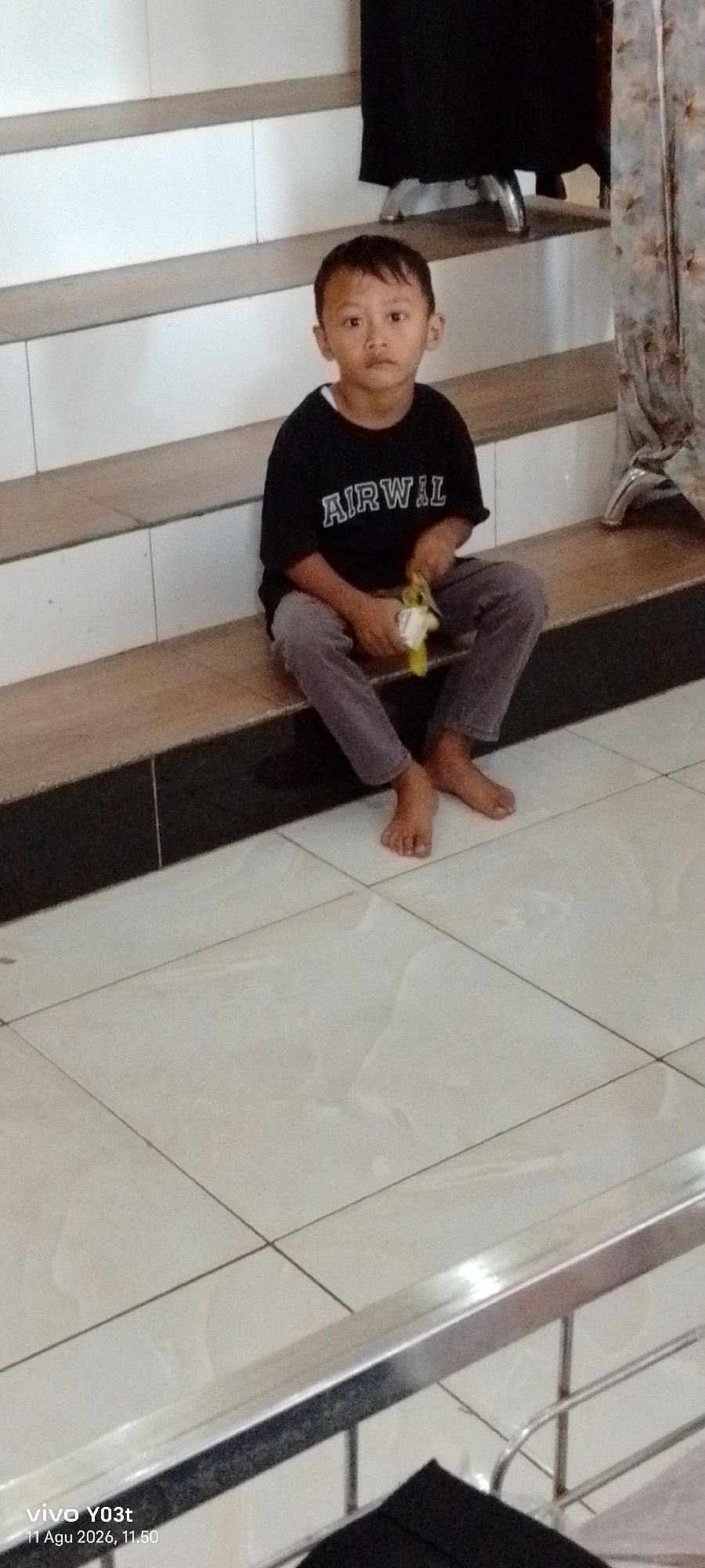 A young boy is sitting on the wooden step of a staircase. He is wearing a black t-shirt with the words "AIRWAL" printed on it in white lettering, and grey pants. The boy is holding a yellow object in his right hand. His feet are bare and he is looking directly at the camera with a neutral expression. The staircase is made of wood with white risers. The floor is tiled with light-colored tiles. Behind the boy, a dark curtain is visible. The overall scene appears to be indoors. This image was taken at 11:50 AM on August 11, 2026, in Atula, Indonesia, as indicated by the timestamp in the lower-left corner of the image.