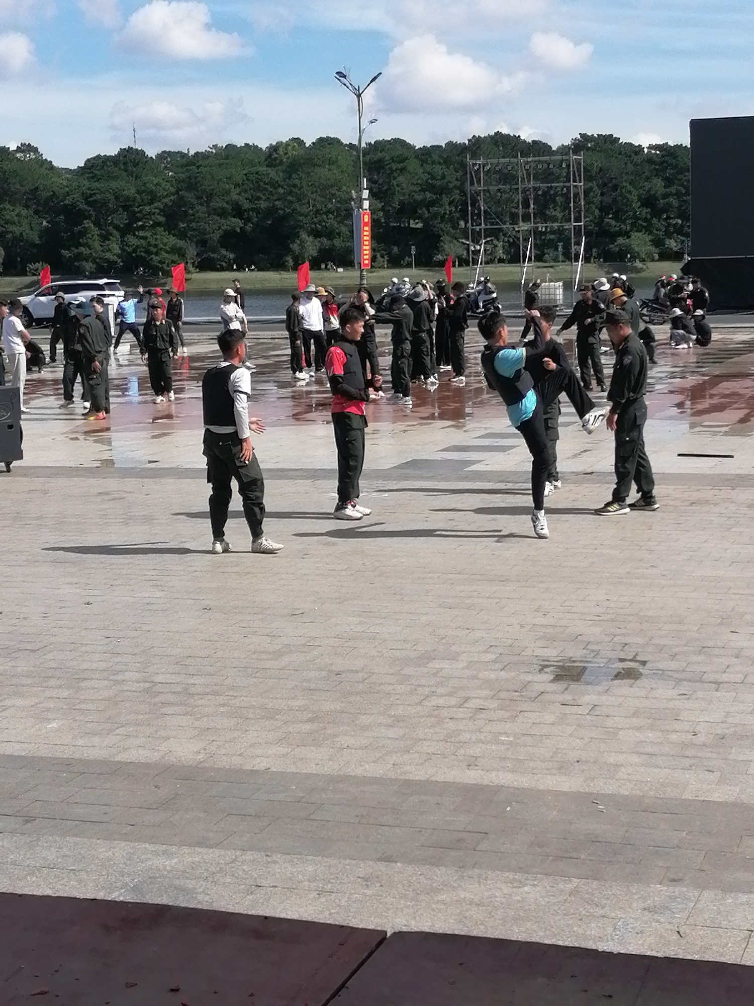 The image depicts a sunny outdoor scene on a paved area, likely a plaza or promenade, with a body of water and trees in the background. Several individuals, mostly young men, are engaged in what appears to be martial arts training or a demonstration. One person in the foreground, wearing a black vest over a white shirt and dark pants, stands facing away from the camera with their right hand extended. To their left, another individual in a blue shirt and black pants is captured mid-kick, demonstrating an athletic maneuver. Further back, groups of people in similar attire are positioned, some observing, others appearing to participate or wait. Red flags are visible, adding a splash of color to the scene. The ground is wet in patches, suggesting recent rain or cleaning. The presence of a white SUV on the left and a large black screen or structure on the right hints at an organized event or performance. The overall atmosphere is active and dynamic. The style of clothing worn by many of the participants, including black vests and dark pants, suggests a uniform or team attire. The environment appears to be a public space in Da Lat, Vietnam, likely near Xuan Huong Lake, a popular tourist destination known for its scenic beauty and recreational activities.