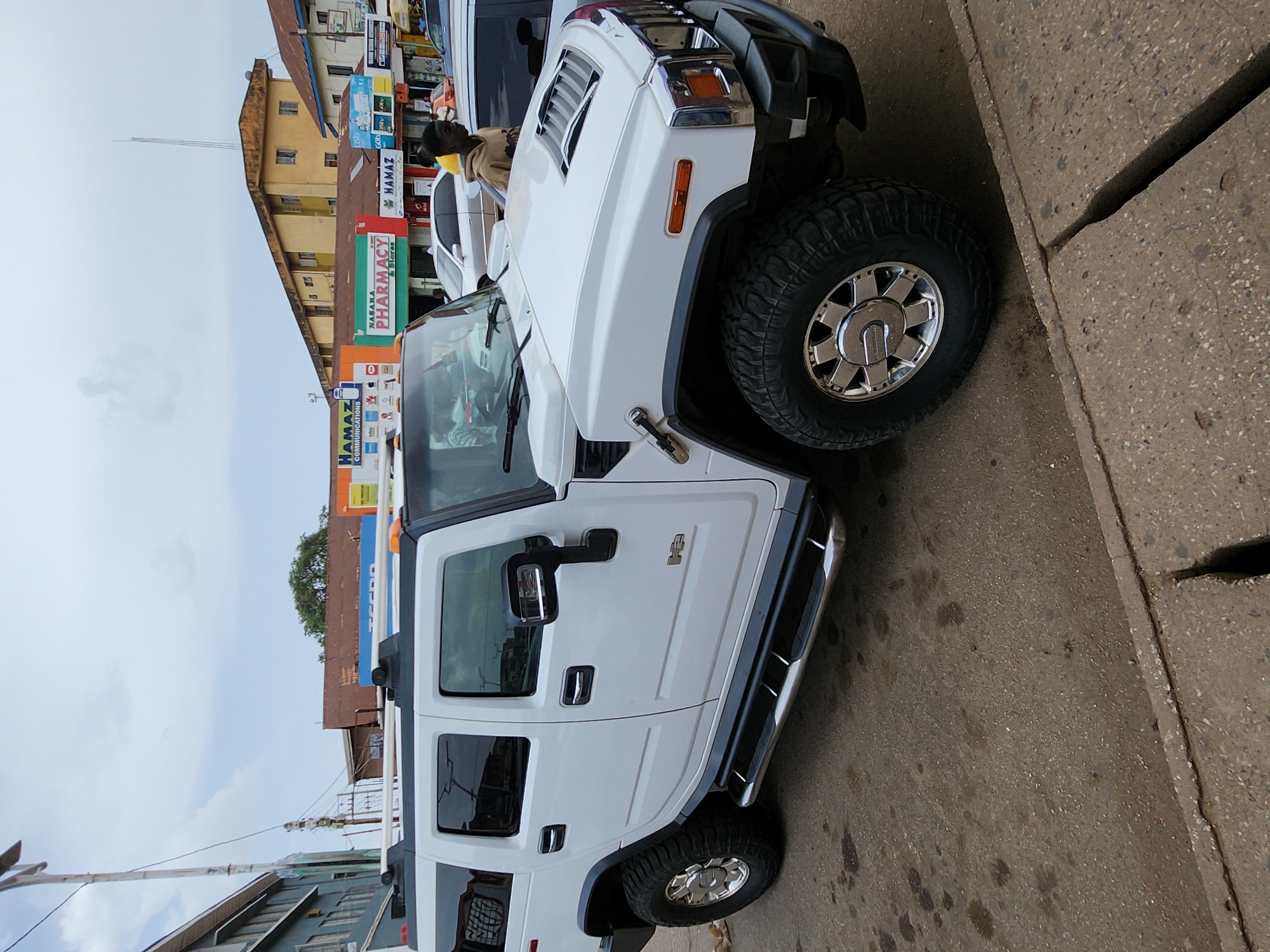 The image captures a white Hummer H2 parked alongside a street in the city of Unknown, Unknown. The Hummer, positioned on the right side of the frame, is partially obstructed by the edge of the sidewalk. Adjacent to the vehicle, the street surface is stained with what appears to be fluid. The background features a line of buildings, some with signs displaying "HAMAZ PHARMACY & Stores" and other smaller advertisements. In the upper portion of the frame, a person is seen partially obscured between two vehicles. The sky is visible above the buildings, revealing a slightly overcast day.