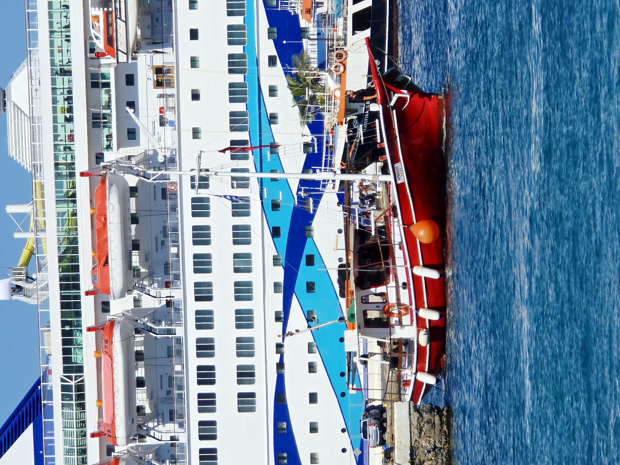The image shows a docked red and white boat in the foreground, with a large white cruise ship in the background. The cruise ship has multiple decks with rows of windows and several lifeboats attached to its side. The boat in the foreground has a wooden structure and is adorned with white buoys and a red life ring. A Greek flag and another flag are visible on the boat's mast. In the middle ground, part of another vessel with an orange stripe is visible. The scene appears to be a harbor in Agios Nikolaos, Greece, with the bright blue water suggesting a sunny day.