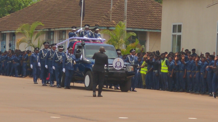 A daytime procession is depicted on a reddish-brown paved road within an apparent official compound in the city of Kilimanjaro, Tanzania. A dark blue SUV, featuring a circular emblem on its front grille, white sidewall tires, and red and blue lights on a roof rack, moves from left to right. Four to five individuals in light blue military-style uniforms with caps and sashes stand in the open rear section of the SUV, appearing to wave or salute. A lone individual in a dark suit or uniform stands directly in front of the vehicle, facing away from the viewer, seemingly guiding the procession. To the immediate left of the SUV, a line of at least six uniformed individuals in matching light blue attire marches. To the right of the road, a large group of individuals in dark blue uniforms and caps lines the route; many are seen raising their right hands in a wave or salute. One individual within this group wears a bright yellow-green reflective vest over their uniform. In the background, a light-colored building with a brown tiled roof and multiple windows is visible on the left, while a lighter-colored building with large rectangular windows stands on the right. Green foliage, including a palm-like plant, is situated between the background structures.