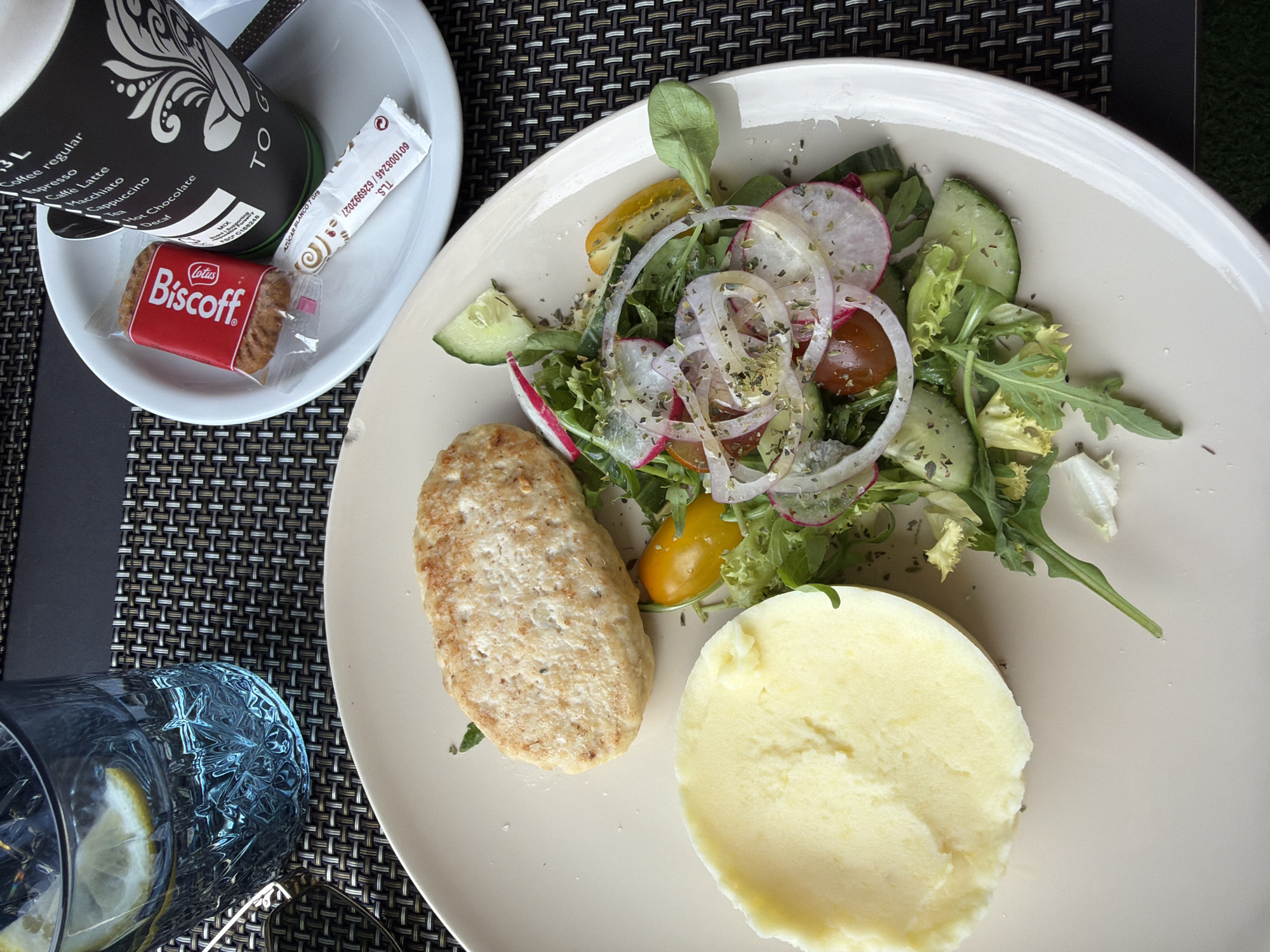 This overhead shot captures a meal served on a textured, woven placemat. The main subjects are a plate of food, a cup, and a glass of water with lemon. The plate holds a small, oval-shaped patty, a mound of mashed potatoes, and a colorful salad. The salad consists of mixed greens, sliced red onions, radishes, cucumbers, and cherry tomatoes, all lightly seasoned. The patty appears to be made of ground meat, possibly chicken or turkey, given its light color and texture. The mashed potatoes are smooth and pale yellow.
Next to the plate is a white cup with a black design, which appears to be a coffee cup with the text "TO GO" prominently displayed. Beside the cup, inside a small white dish, are a single wrapped Biscoff cookie and a sugar packet. The sugar packet has some text and a swirled pattern.
To the left of the plate, a partially visible glass is filled with water and lemon slices. The glass has an intricate, textured design.
The setting suggests an outdoor meal, possibly at a cafe or restaurant, given the placemat and the overall presentation. The lighting is bright, indicating daytime, and there are no visible people. The food is presented neatly, suggesting a light meal or snack. The visible text includes brand names like "Lotus Biscoff" and functional labels on the coffee cup and sugar packet. The scene is peaceful and focused solely on the food and accompanying items.