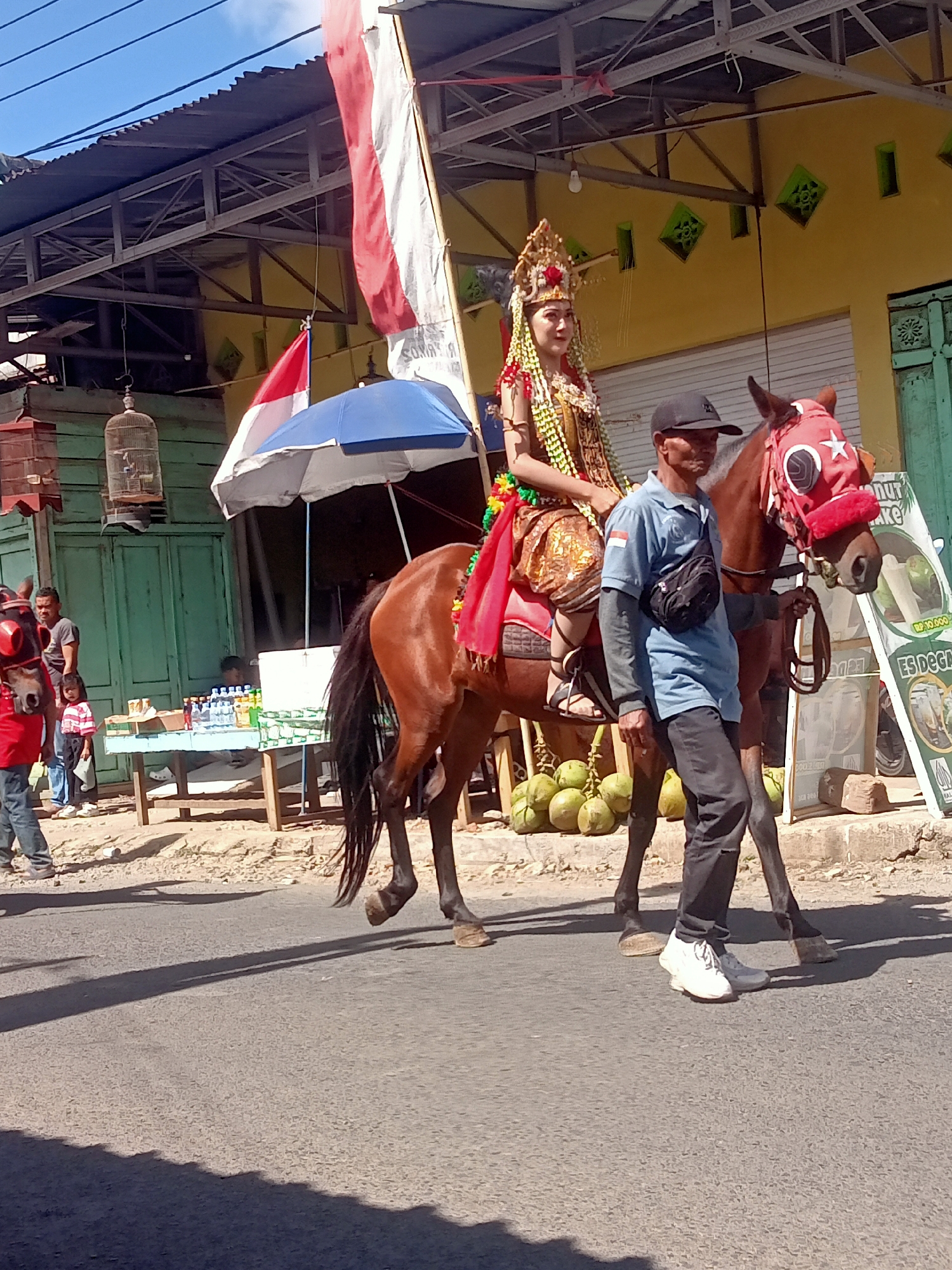 A brown horse is being led down a paved street in Pule, Indonesia. A young woman wearing traditional Indonesian attire, including a headdress, sits on the horse. A man in a blue shirt and black pants walks beside the horse, holding its bridle. In the background, a yellow building with green trim and a corrugated metal roof is visible. There are also several coconuts on the ground and a sign for "ES DEGA." The sky is blue with some clouds.
