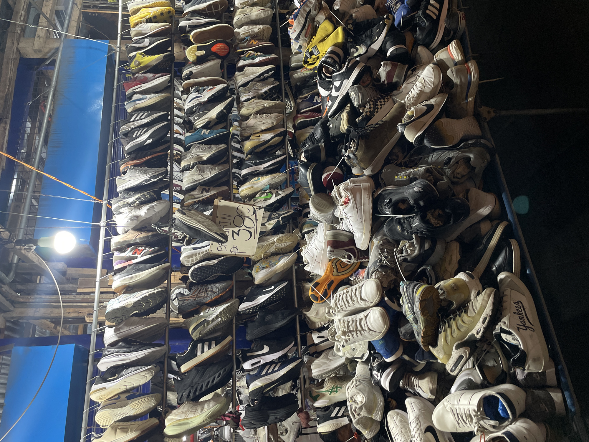 This image depicts an outdoor stall selling a large assortment of athletic shoes, likely in a street market setting in Davao City, Philippines. The shoes are displayed on multiple racks and piled on shelves. The stall appears to be open-air, with a metal roof or awning overhead and what looks like wooden structural elements and blue plastic sheeting in the background. A bright lightbulb hangs above, illuminating the scene. The shoes are of various brands, styles, and colors, suggesting a mix of new and used items. Some shoes are neatly hung on hangers, while others are stacked haphazardly. There are no people clearly visible in the image, but the presence of the stall and the merchandise indicates commercial activity. The overall impression is of a bustling marketplace where a wide selection of footwear is available for purchase.