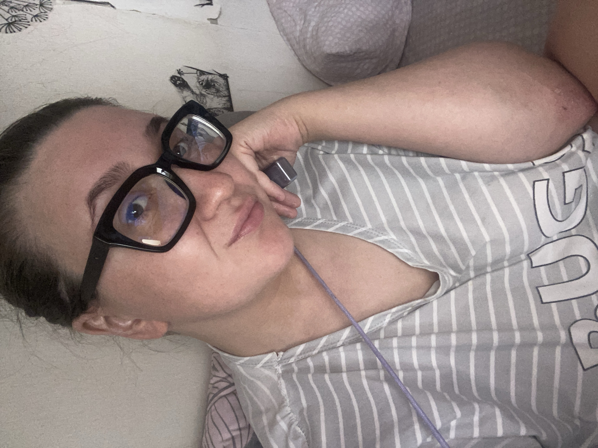 A woman lies on a bed, wearing large, black-framed glasses and a grey and white striped shirt. A grey object rests near her hand. A light purple cord runs from her shirt towards her chin. Behind her, the headboard is decorated with a cat design. The location is unknown but possibly Russia, as that's what the user requested.