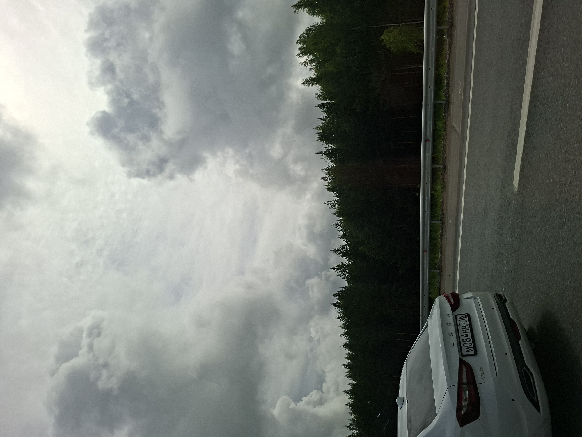 A white Lada Vesta sedan with a Russian license plate (M084HH 716) drives on a highway. The car is seen from the rear passenger side. The highway is bordered by a dense forest of dark green trees and a metal guardrail. The sky is heavily overcast with dramatic grey clouds, suggesting impending rain or a stormy weather condition. The image appears to be taken from another vehicle moving on the same highway, giving a dynamic perspective of travel. The overall scene suggests a journey through a natural, possibly remote, area. The location is identified as the city of Pokrovka, Russia.