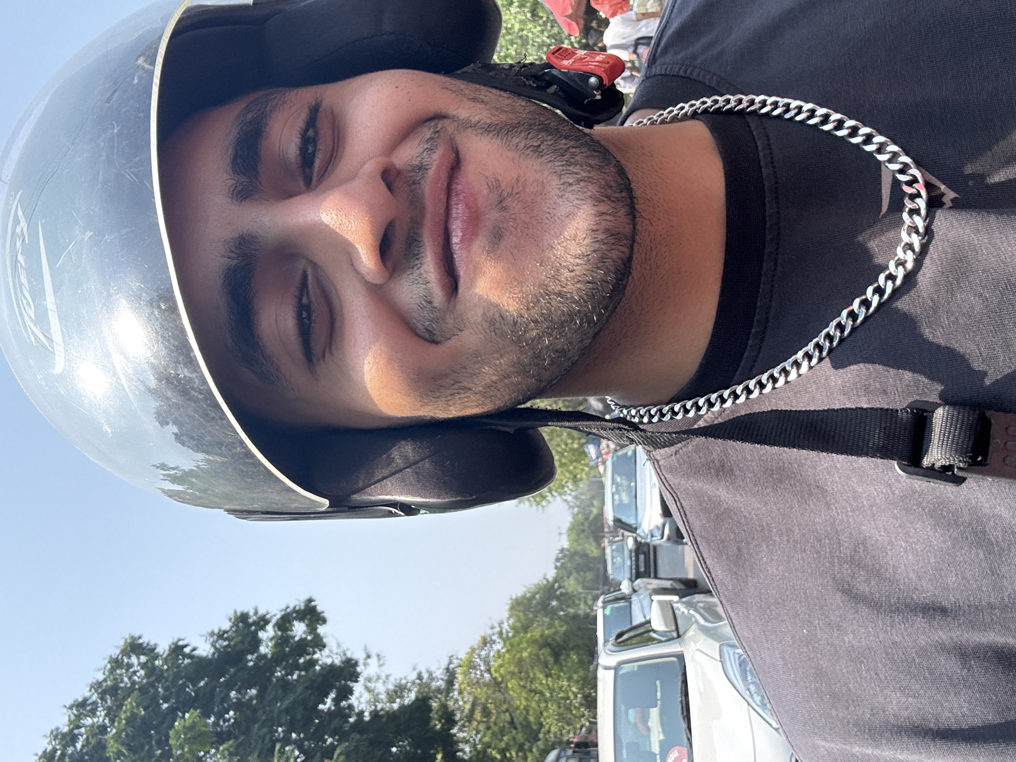 A person wearing a helmet and a silver chain stands in what appears to be a traffic jam. The background reveals trees and a clear sky, which suggests the scene is taking place outdoors during the day. Several vehicles are visible behind the person. The person looks toward the camera with a slight smile. Kolkata, India is not inferable.