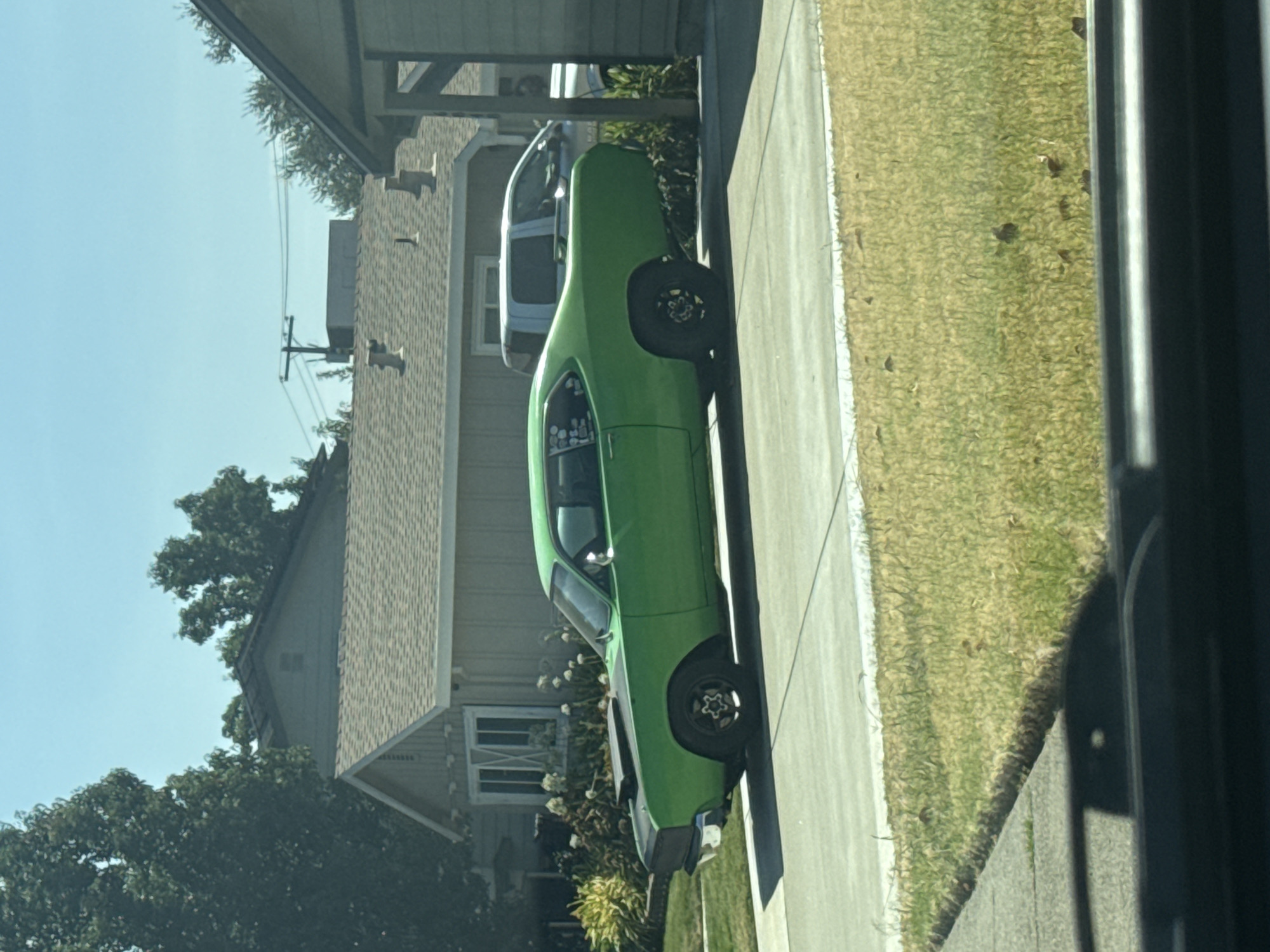 A bright green vintage muscle car with a black hood scoop is parked on a paved driveway. The car has a custom look, with a lowered stance and aftermarket wheels. In the background, a light blue pickup truck is parked in a driveway next to a suburban house with a beige exterior and a brown shingled roof. The scene appears to be in a residential neighborhood in Citrus Heights, California, United States of America, under a clear blue sky.