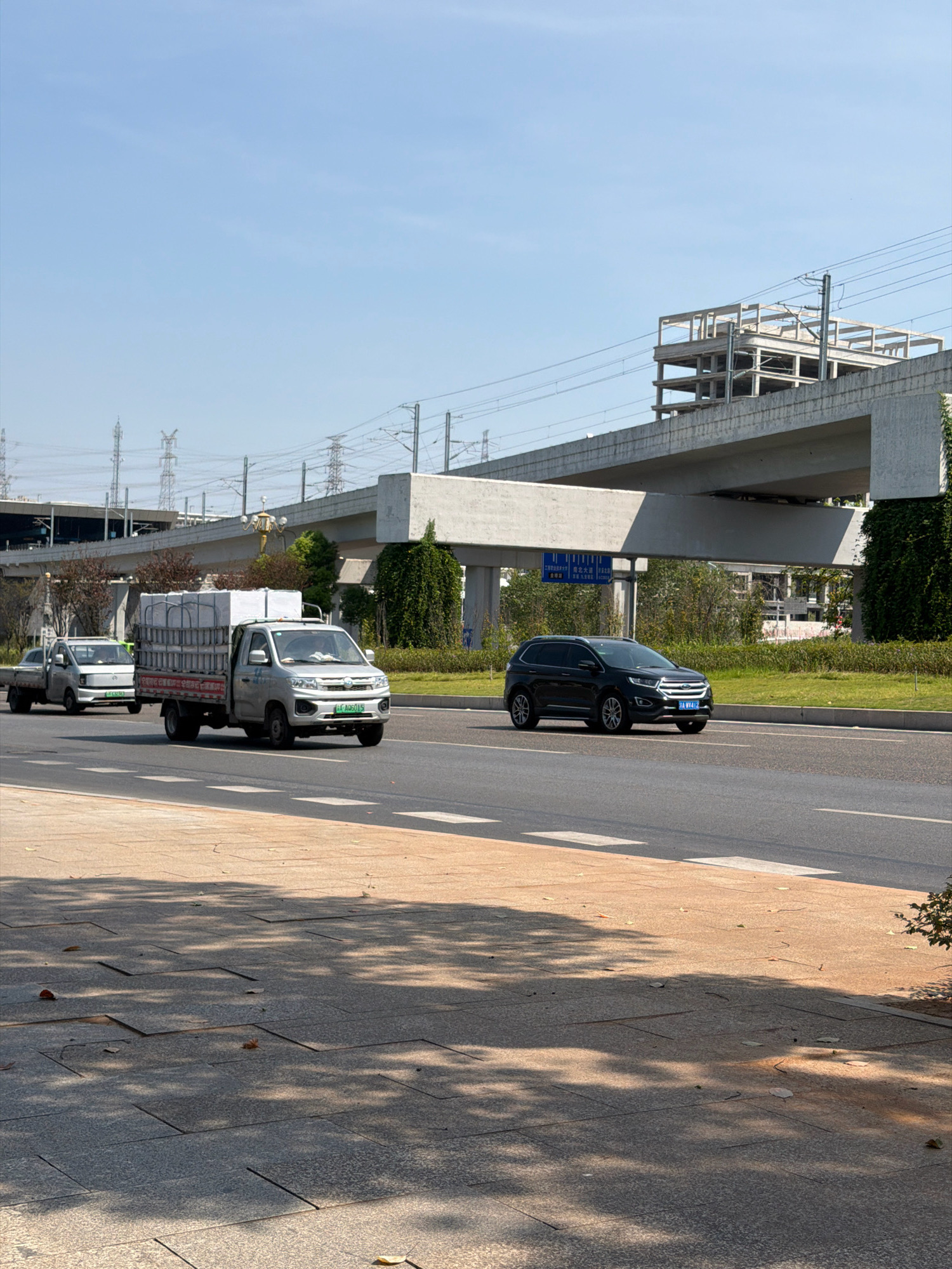 This is an outdoor, eye-level, medium shot of a street scene in Jiangjin, China, under a clear, bright sky. A multi-lane road is the primary focus, with several vehicles present. In the foreground, a paved sidewalk with scattered leaves and dappled sunlight from overhead foliage runs horizontally across the frame.
On the road, from left to right, a light-colored, small truck with a cargo bed filled with white boxes is traveling away from the viewer. To its left, another similar, smaller light-colored truck is also moving in the same direction. Further to the right, a dark-colored SUV is traveling towards the viewer. The road has white lane markings.
Overhead, a large concrete overpass with a skeletal concrete structure under construction on top runs across the mid-ground of the image. Beyond the overpass and to the left, power lines and pylons are visible, suggesting an industrial or transportation hub. Green grass and some trees line the roadside next to the overpass. A blue and white street sign is affixed to a pole underneath the overpass. The overall scene suggests a developed urban or suburban area with ongoing infrastructure projects.