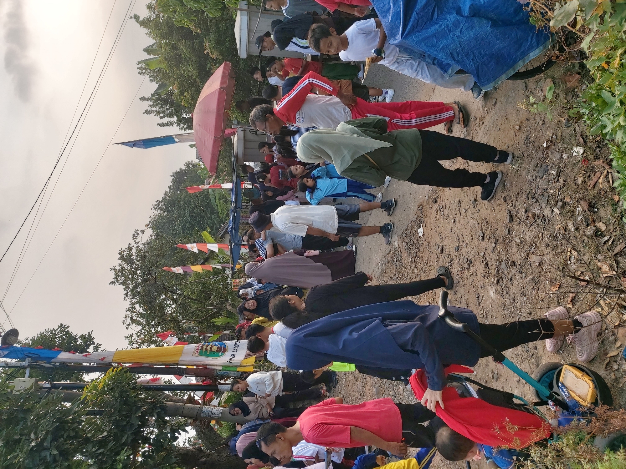 This image captures a lively outdoor gathering in Bandar Lampung, Indonesia, likely a community event or celebration, evidenced by the presence of numerous people of various ages and the festive decorations. In the foreground, a person in a blue hijab pushes a stroller containing a child, with a red blanket draped over it. The ground is a mix of dirt and small rocks, with scattered dry leaves and low-lying vegetation.
A diverse crowd populates the scene. Several individuals are dressed in casual attire, while others wear more athletic wear, suggesting potential recreational activities. Some are seen conversing, while others appear to be observing or participating in the event. A man in a red and white tracksuit stands out, looking towards the right of the frame. A red umbrella provides shade in the background, near a fence.
Decorations are visible throughout the image. Flags, including the Indonesian flag (red and white) and other colorful banners, are strung up along poles and wires, indicating a festive atmosphere. One prominent flag displays the emblem of Bandar Lampung, further pinpointing the location. Trees with lush green foliage line the background, contrasting with the overcast sky, which suggests it might be early morning or late afternoon. The overall impression is one of a community coming together for a shared occasion.
