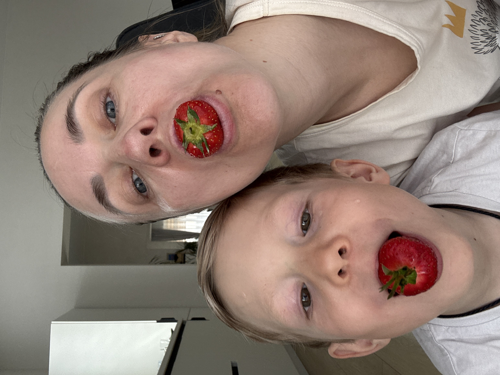 A headshot photo, rotated 90 degrees counterclockwise, shows a woman and a boy with strawberries in their mouths. The woman is on top, wearing a light-colored shirt with a crown and bird design. The boy is beneath her, wearing a white shirt with a black collar trim. Both individuals are looking directly at the camera. The background consists of white walls and a white cabinet. There is no definitive evidence to place the image in Chaikovsky, Russia.