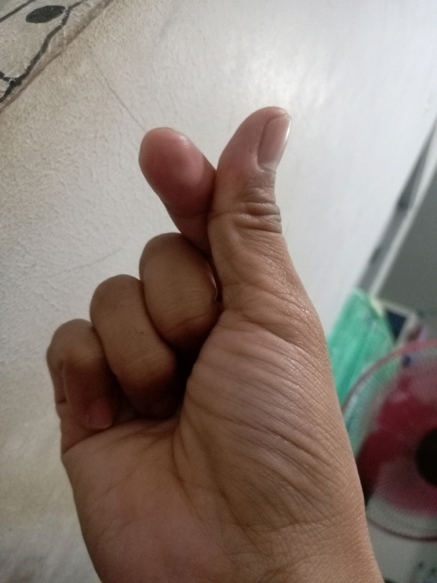 A close-up shot shows a human hand forming a heart shape with the thumb and index finger. The hand is positioned in the foreground, with the fingers of the left hand curled into the palm. The background is blurred, but appears to be an indoor setting with a textured wall and some out-of-focus objects that suggest a domestic environment. The lighting is soft and natural. There is no information about the location in Batam, Indonesia.