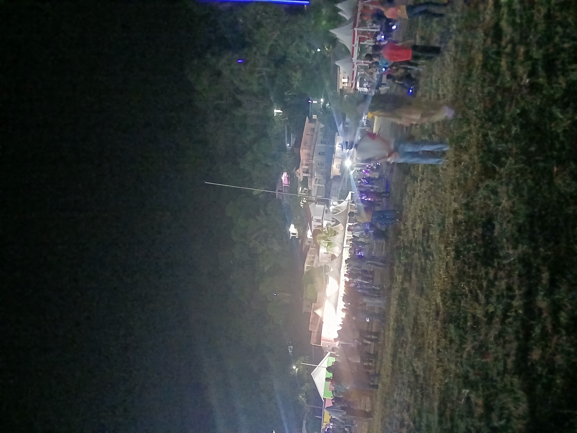 The image depicts an outdoor nighttime gathering at what appears to be a community event or festival. The scene is characterized by low-light conditions, with artificial lighting illuminating a crowd of people milling around. In the background, a hillside is visible with several buildings, some of which are illuminated, suggesting a residential or commercial area situated on the slopes. Tents are erected in the foreground, indicating stalls or activity areas typical of such events. Several individuals are visible in the frame, some standing and observing, while others appear to be walking through the open space. The ground is covered in grass, and there is a slight haze or fog present, possibly due to smoke from food vendors or atmospheric conditions. The overall impression is one of a lively, albeit dimly lit, social gathering taking place in a semi-urban or semi-rural environment.