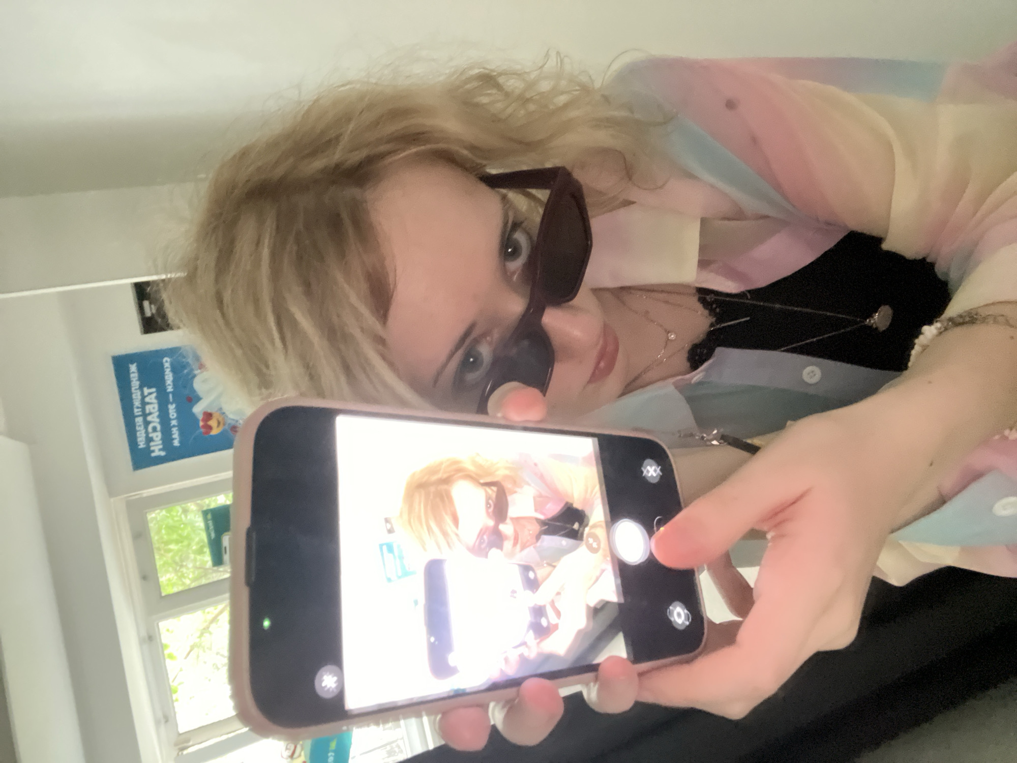A young woman with blonde hair and sunglasses is taking a selfie with a smartphone in her left hand. She is wearing a multicolored shirt and a black camisole with a lace trim. The phone screen shows a reflection of the woman, who appears to be in an indoor setting with a window in the background that looks out onto trees. There is a poster on the wall behind her with text that appears to be in Cyrillic script. The overall lighting suggests it is daytime. There is no discernible location within Almaty, Kazakhstan.