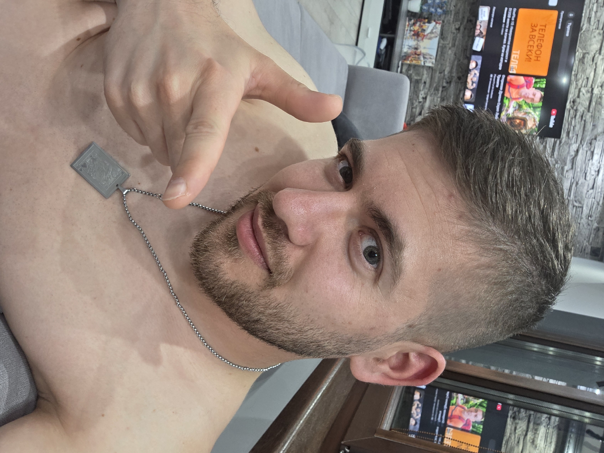 A man with short, brown hair and a stubbly beard is posing for a selfie. He is shirtless and wearing a silver chain necklace with a square pendant that appears to be a playing card symbol. His right hand is raised, with his index finger pointing towards the camera. The background shows a grey couch, a TV displaying content in another language, and a partially visible window. There is no information about the location of Yagodovo, Bulgaria in the image.