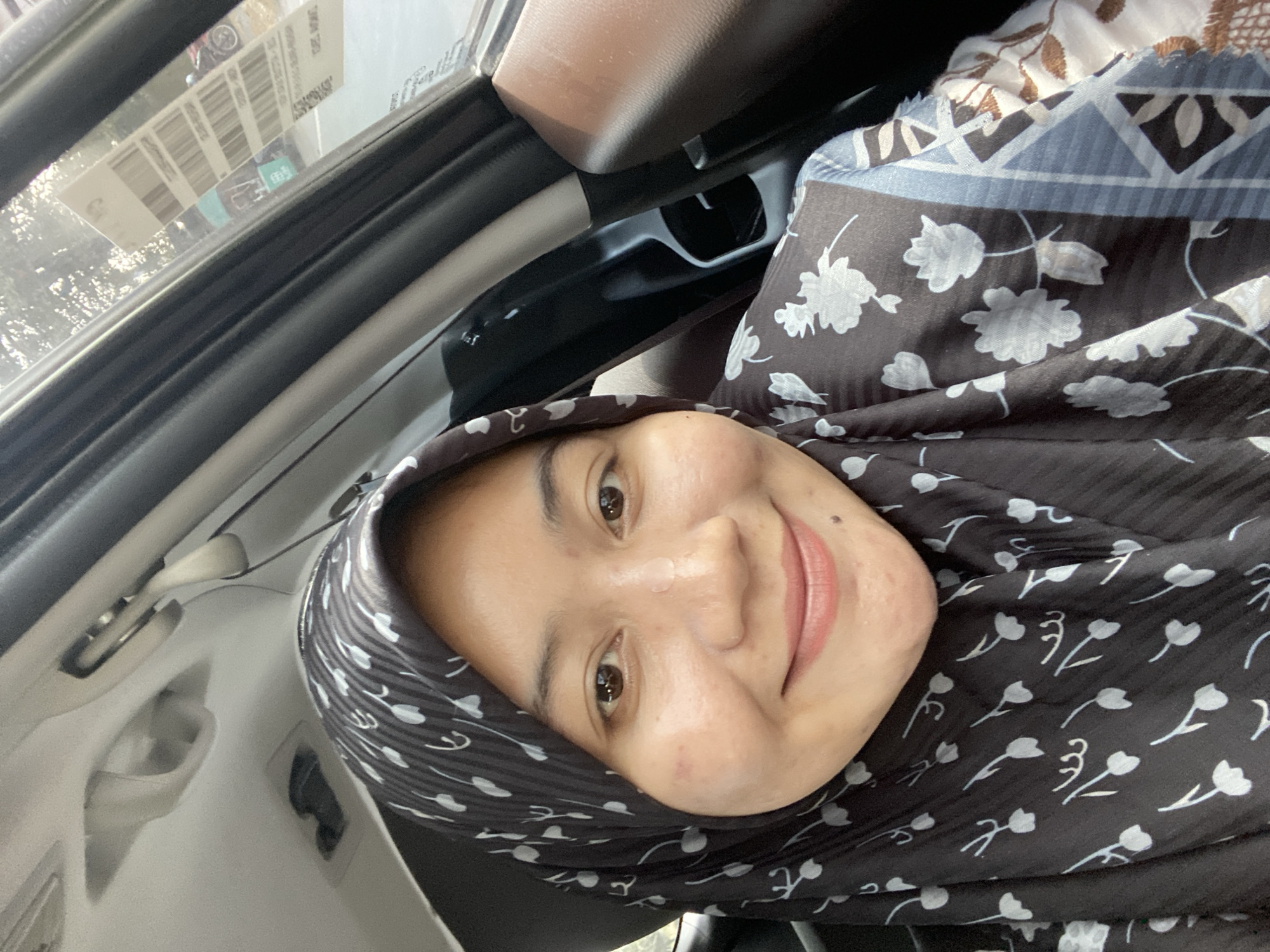 A young Indonesian woman wearing a black hijab with a white floral pattern and a light blue and white patterned garment is pictured inside a car. She is smiling and looking towards the camera. The interior of the car is visible, including the roof lining, a grab handle, and a portion of the passenger seat. Outside the car window, sunlight filters through trees, and a sticker with barcodes and text is affixed to the glass. The scene appears to be an outdoor setting, potentially a street or parking area within Banjarbaru, Indonesia.