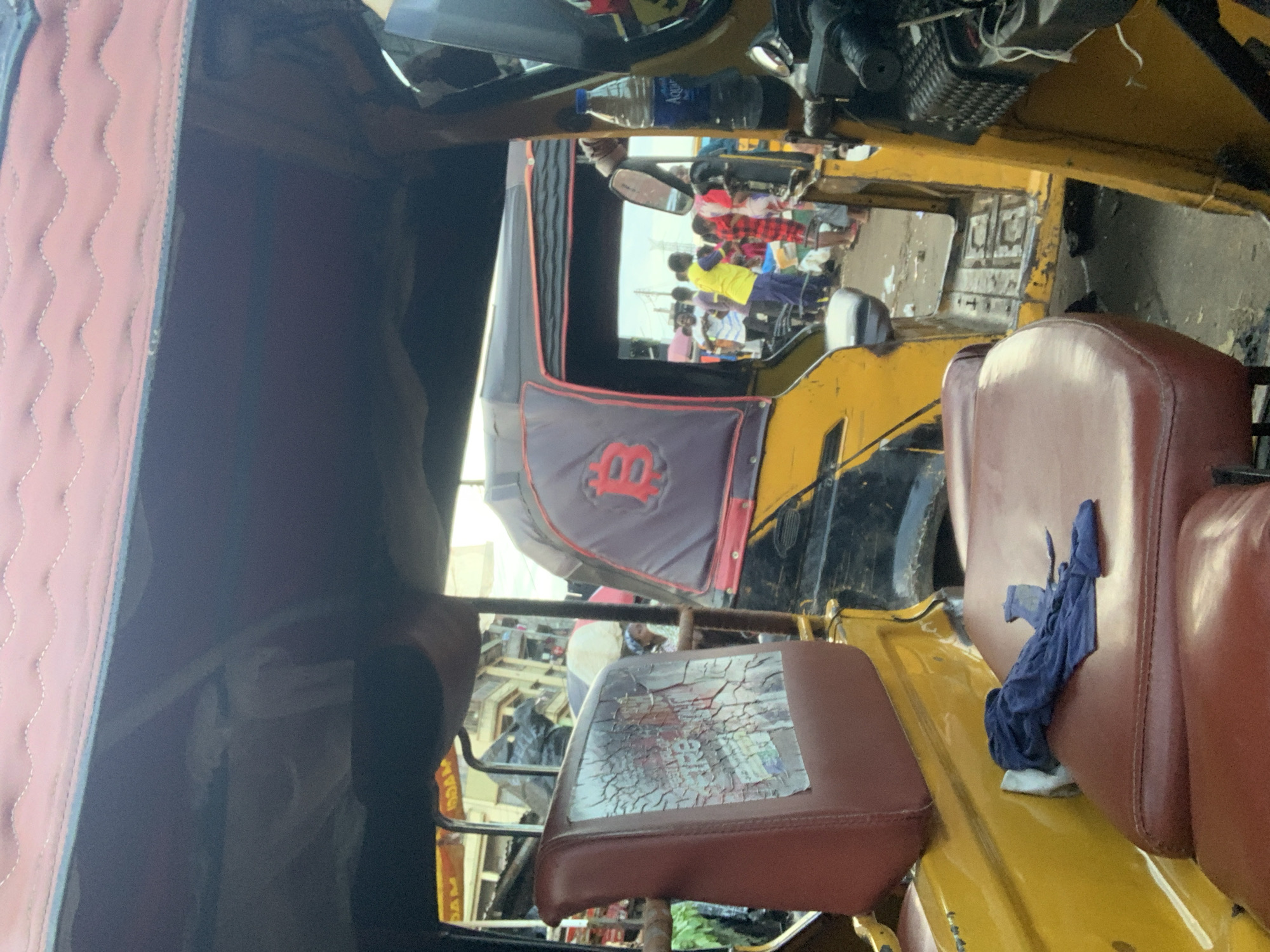 This image depicts the interior of a yellow and black auto-rickshaw, possibly in Obosi, Nigeria, a known hub for auto parts and manufacturing. The auto-rickshaw is parked on a street with a bustling environment visible through the open side. The interior features worn, red leather seats and a dark fabric canopy overhead. A blue cloth is draped over one of the seats. The exterior of the auto-rickshaw is visible through the opening, showing a prominent red Bitcoin logo on the side of another vehicle. Several people, including men and women in casual attire, are seen in the background, some appearing to be shoppers or vendors. The ground is paved and shows signs of wear and tear, with scattered debris. The scene suggests a typical street-side scene in a busy commercial area.
