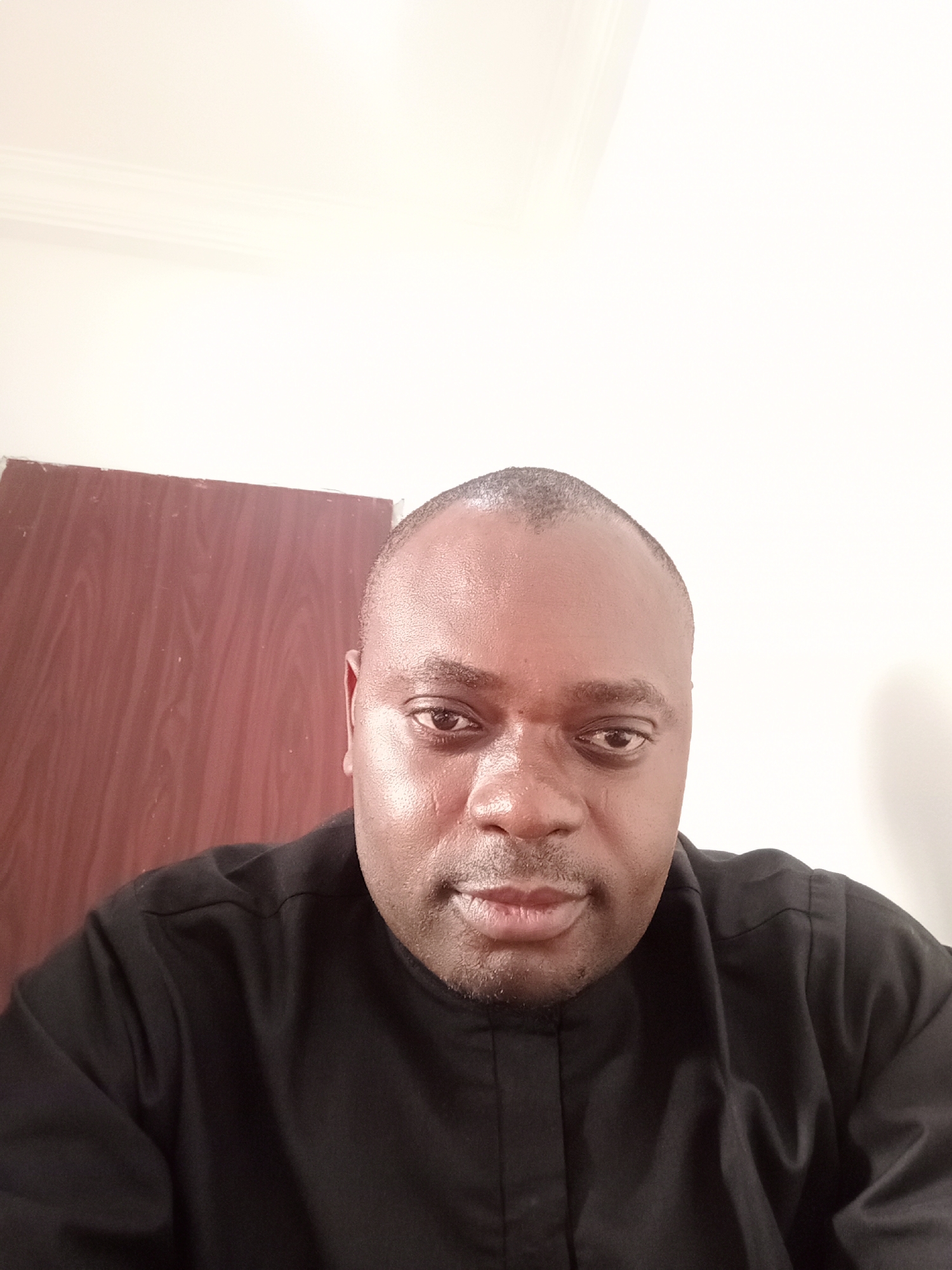 A man with dark skin is centered in a close-up portrait, looking directly at the camera with a neutral expression. He has a shaved head or very short hair and minimal facial hair around his chin and mouth. His skin appears somewhat shiny, with highlights on his forehead and nose. He is wearing a black collared top, possibly a traditional or formal shirt.
The background on the left features a brown wooden door or panel. The remainder of the background is a plain, light-colored wall, possibly white or off-white. A subtle architectural detail, resembling crown molding, is visible in the top left corner where the wall meets the ceiling. The lighting is bright and even, suggesting an indoor setting. No specific activity is taking place other than posing for the photograph. There is no visible text in the image. The setting is indoors, with the location context being Gwarinpa, Nigeria.